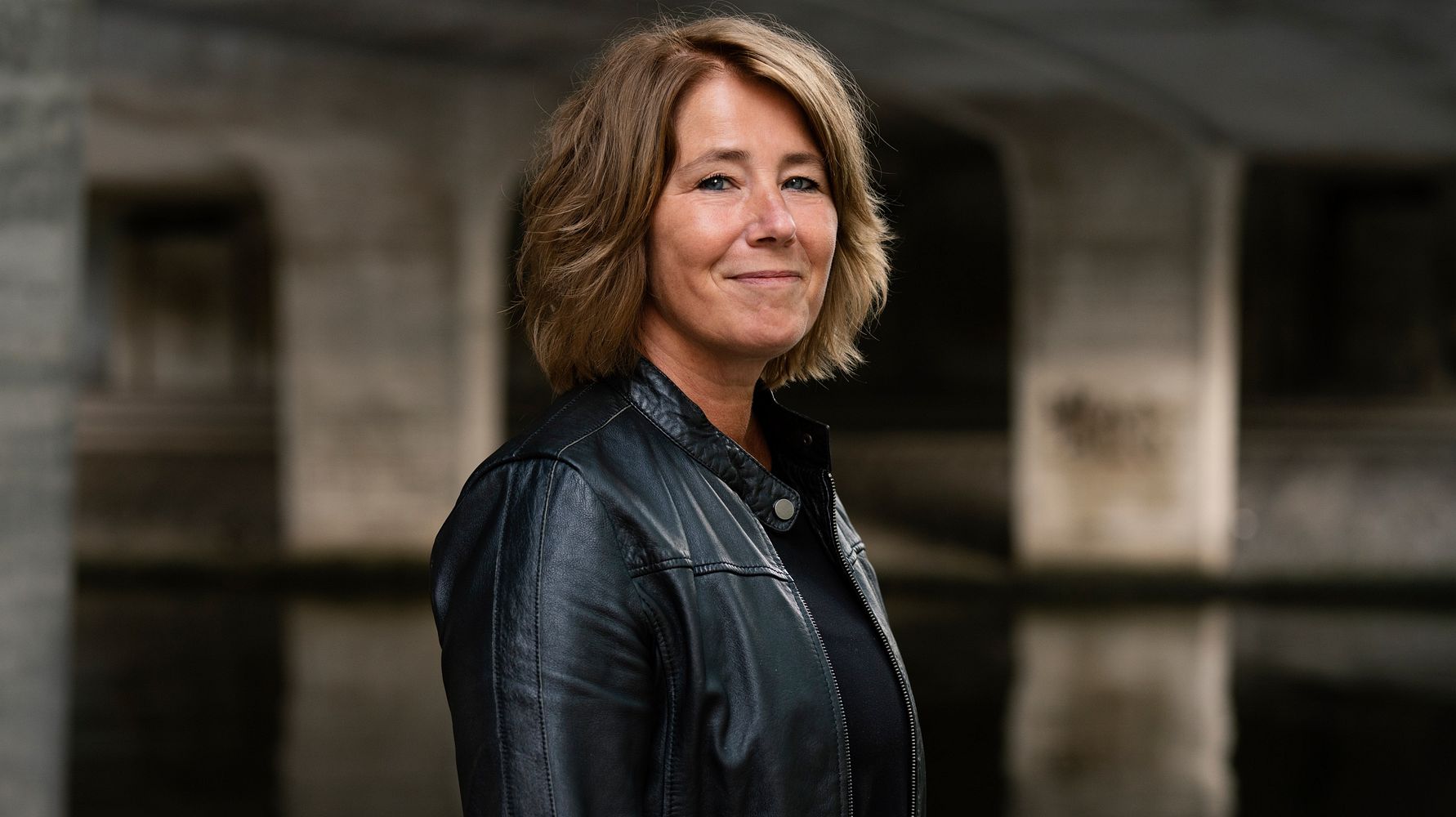 Person med kort, blont hår klädd i en svart läderjacka, står framför en bakgrund med betongstrukturer och vatten