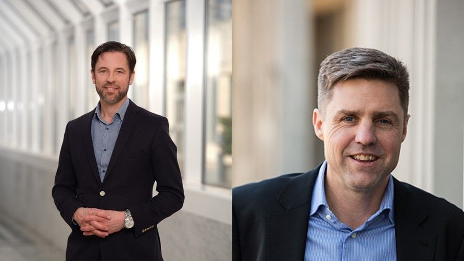 Man klädd i mörk kavaj och blå skjorta står inomhus i en ljus korridor med stora fönster