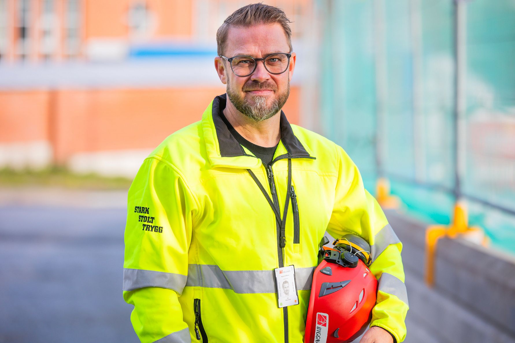 En person i gul reflexjacka som håller en röd hjälm, står utomhus nära en byggnad och vägavspärrning