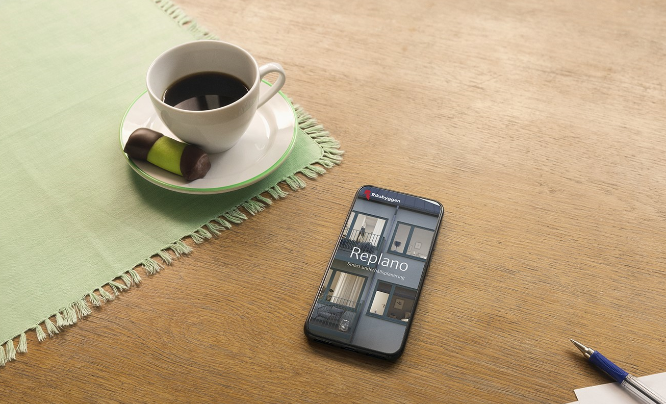En smartphone med appen Replano visas på skärmen, placerad på ett träbord bredvid en kaffekopp, en grön servett, choklad och en penna