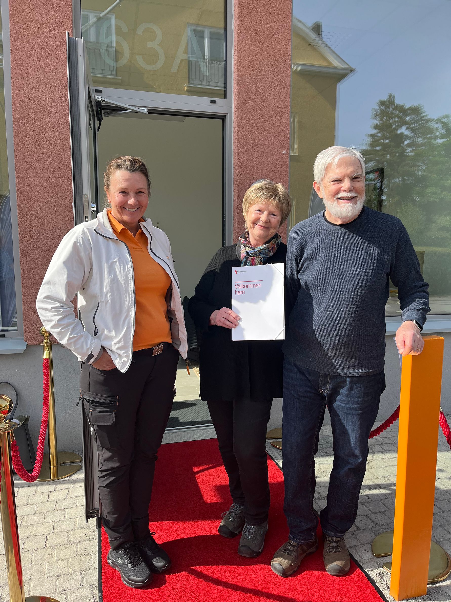 Tre personer står på en röd matta framför en glasdörr med ett rött rep på sidorna, en av dem håller ett dokument i handen
