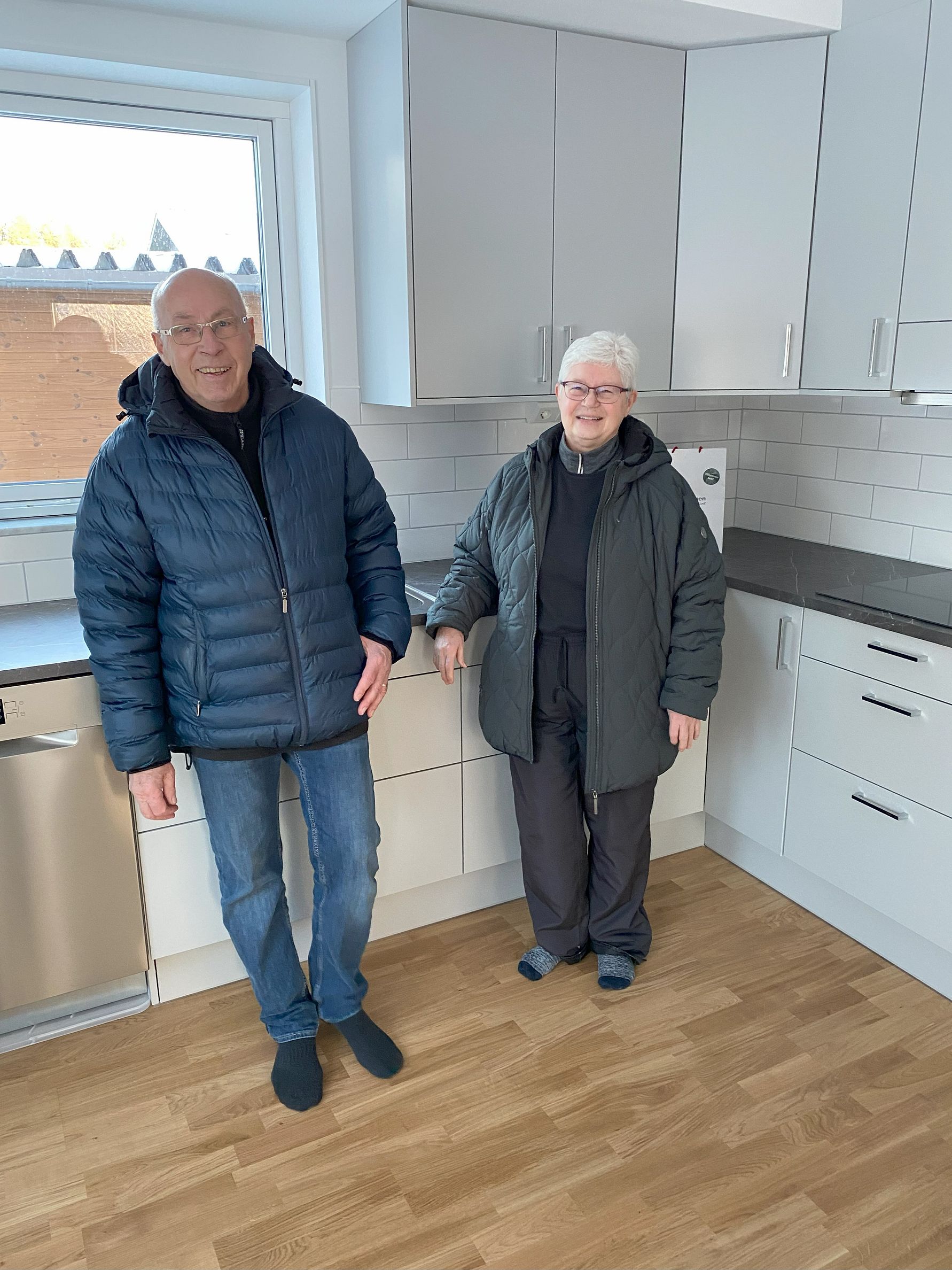 Två personer står i ett ljust kök med vita skåp, trägolv och kaklade väggar; den ena personen har på sig en mörkblå jacka och jeans, medan den andra bär en grå jacka och mörka byxor