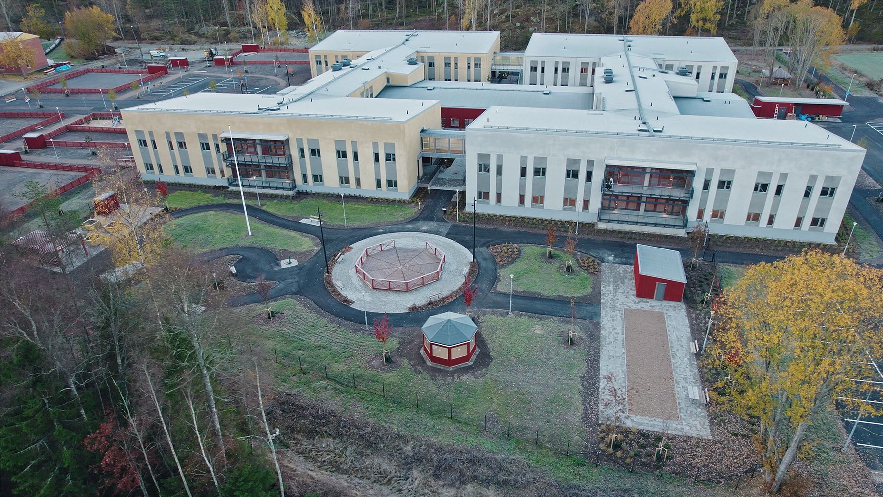 Flygbild av en modern skolbyggnad med vit fasad, omgiven av grönområden och höstfärgade träd. En lekplats med ett cirkulärt område och paviljong syns i förgrunden