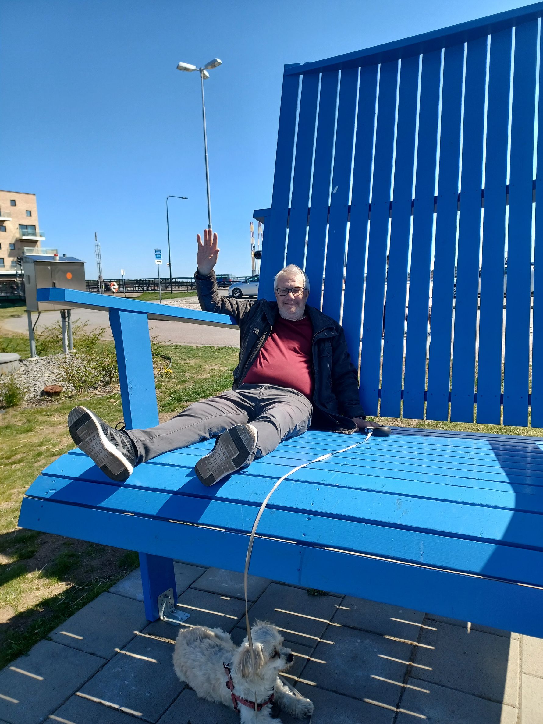 Man sitter avslappnat på en stor blå bänk utomhus med en liten hund kopplad vid sina fötter. Bakgrunden visar en byggnad, grönska och en klar blå himmel