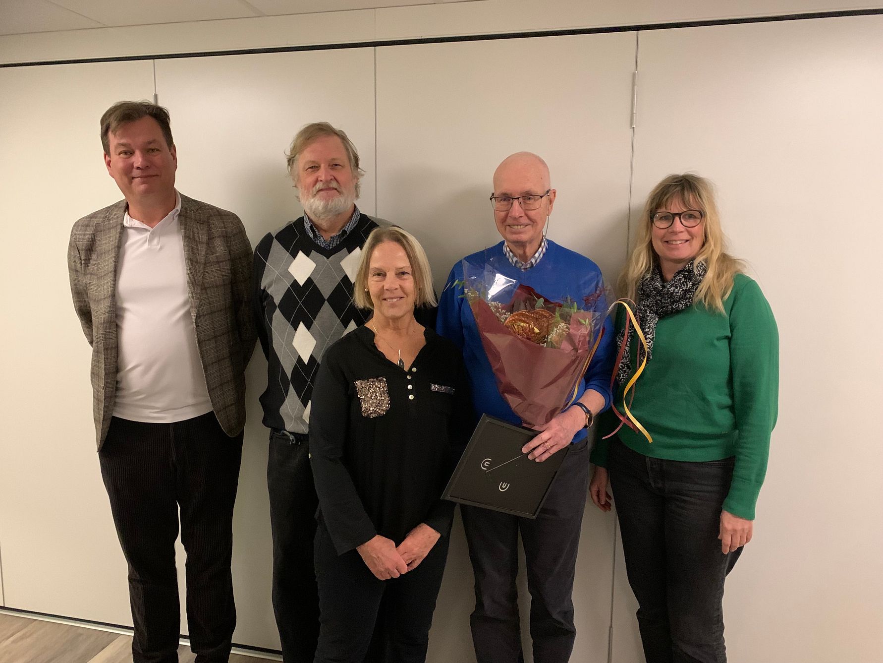 En grupp personer står framför en vit vägg, en person håller en blombukett och ett diplom