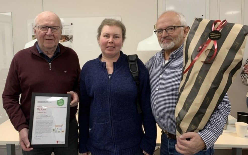 Tre personer står tillsammans inomhus. En person håller i ett diplom, och en annan håller en blombukett inslagen i randigt papper med ett rött band