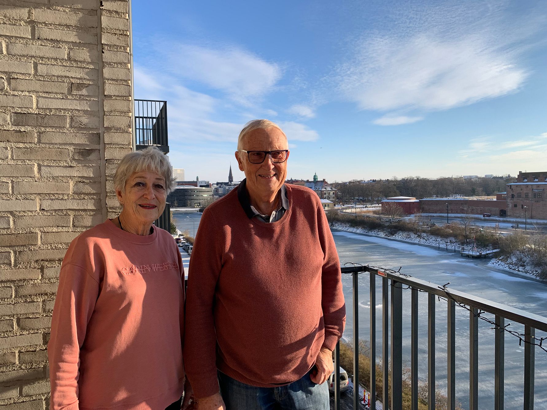 Två personer står på en balkong med en flod och byggnader i bakgrunden under en klarblå himmel