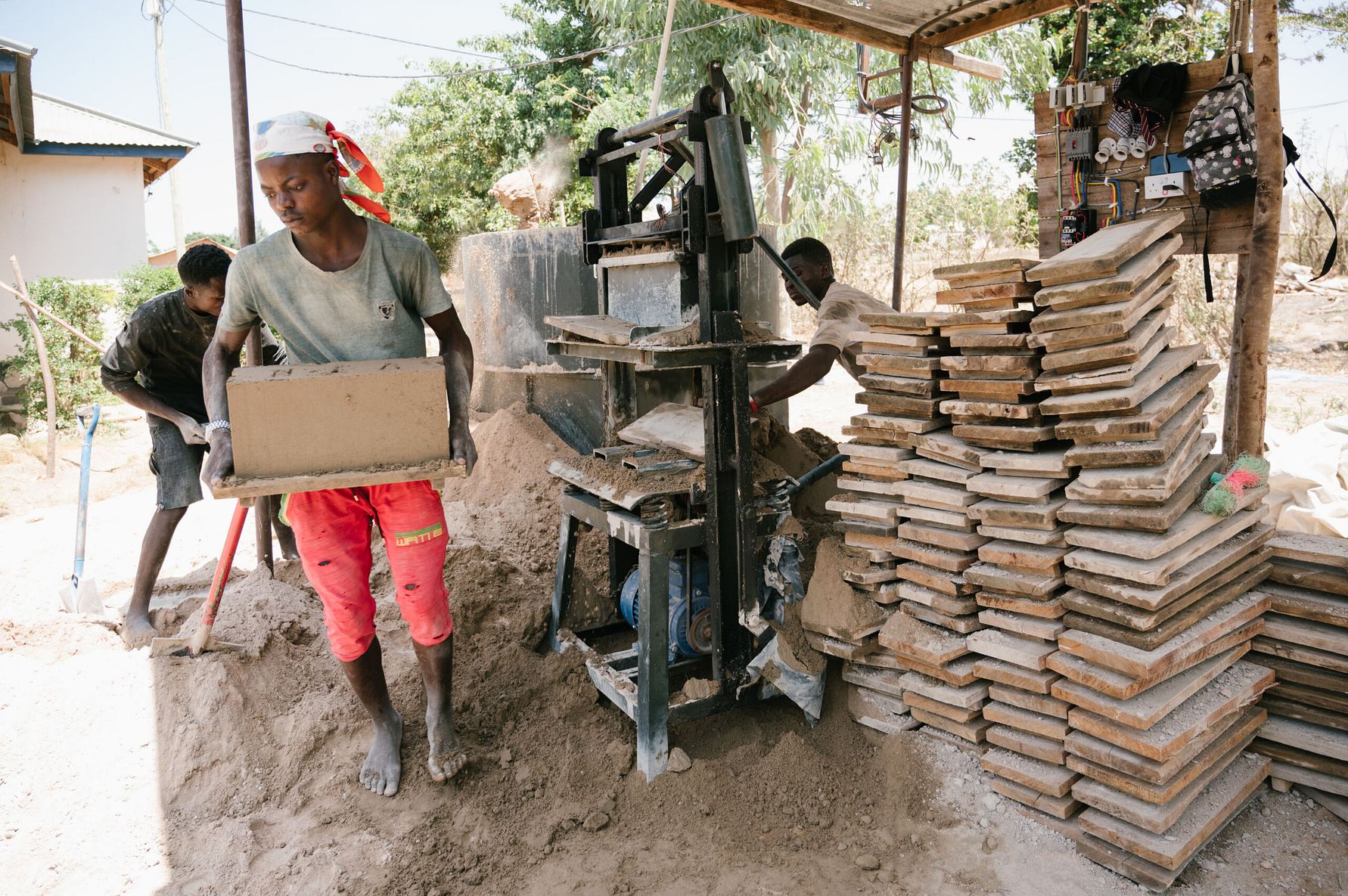 En man i grå t-shirt och röda shorts bär en tegelsten i en utomhusmiljö vid en maskin för tegelproduktion, omgiven av staplade tegelstenar och sand