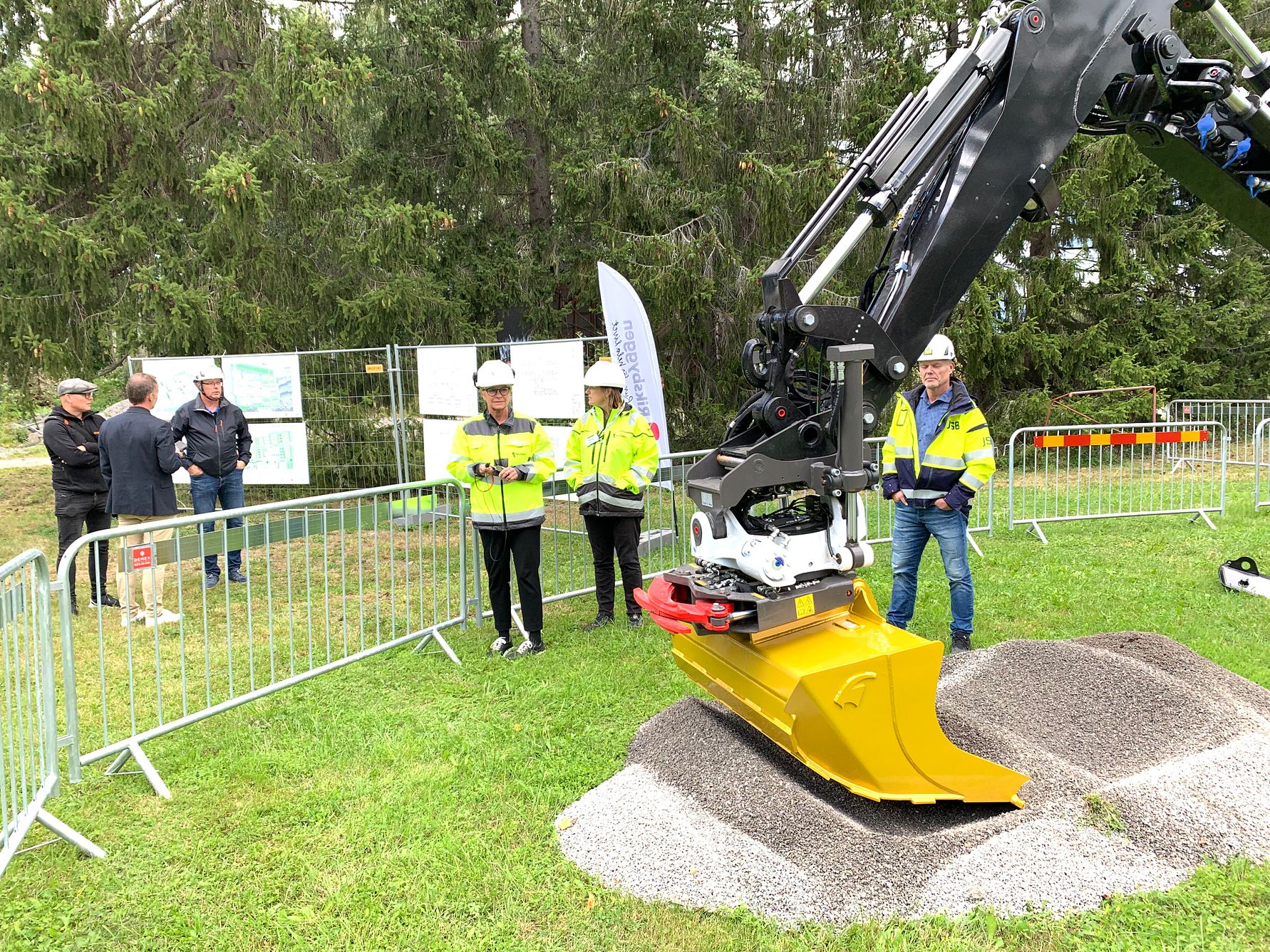 Personer i skyddskläder står intill en stor grävmaskin med ett gult skopliknande verktyg, omgivna av gräs och träd samt avspärrningar och informationsskyltar i bakgrunden