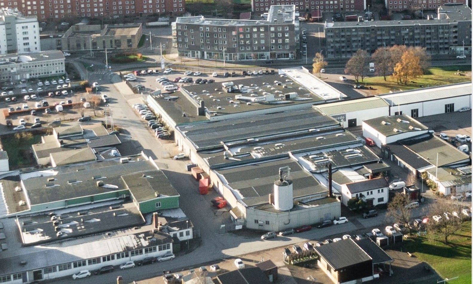 Flygvy över ett industriområde med flera byggnader, parkeringsplatser och omgivande bostadshus