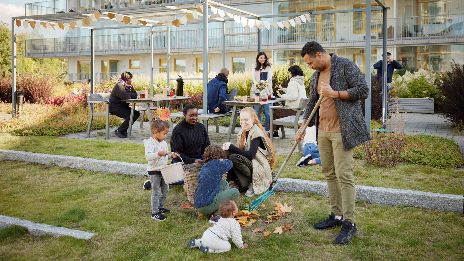 Familj och grannar som samlas på en gräsmatta för att kratta löv och umgås, med en modern byggnad och sittgrupper i bakgrunden