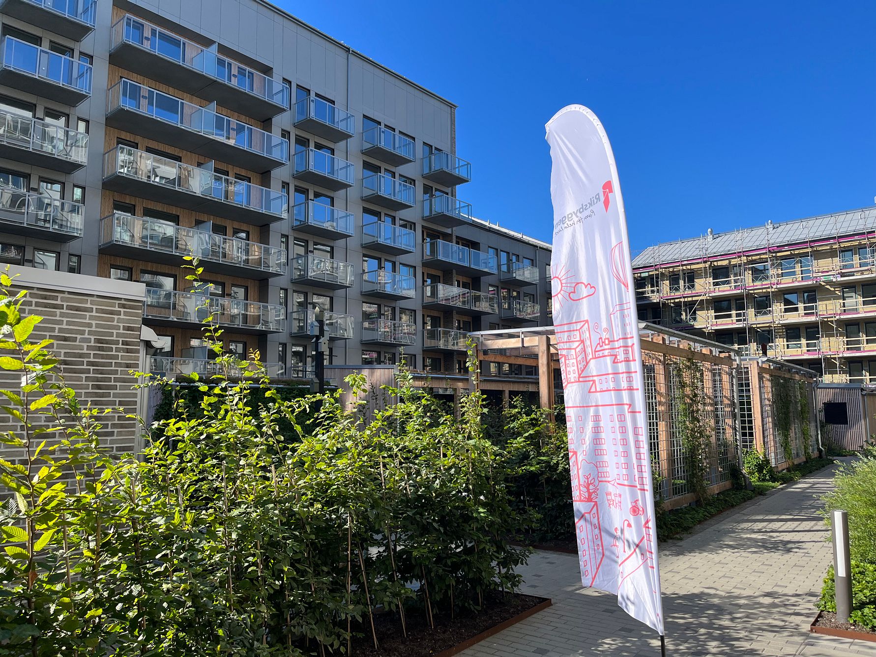 Flerbostadshus med moderna fasader och balkonger, omgivna av grönska och en flagga med tryck i förgrunden under en klarblå himmel