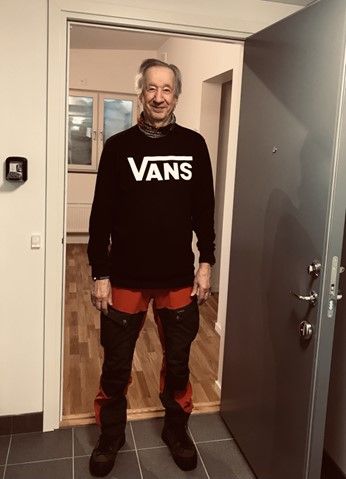 En person i svart Vans-tröja och röda byxor med svarta knäskydd, står i en dörröppning med ett golv av grå och träfärgade material i bakgrunden