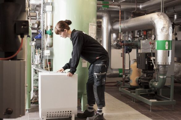 Person arbetar i ett tekniskt rum med rör, ventiler och maskiner, stående vid en vit maskin