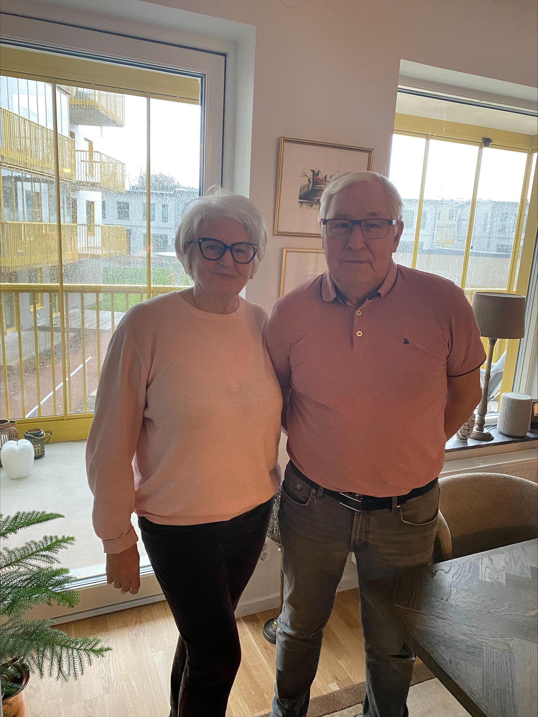 Två personer står i ett rum med stora fönster som vetter ut mot en balkong. I bakgrunden syns gulmålad balkongräcke och flerbostadshus