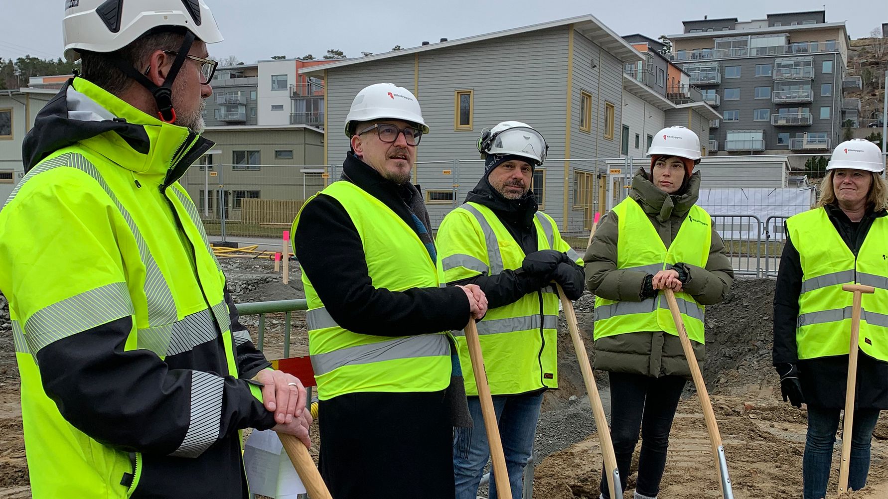 Grupp av personer i reflexvästar och hjälmar håller i spadar vid en byggarbetsplats med bostadshus i bakgrunden