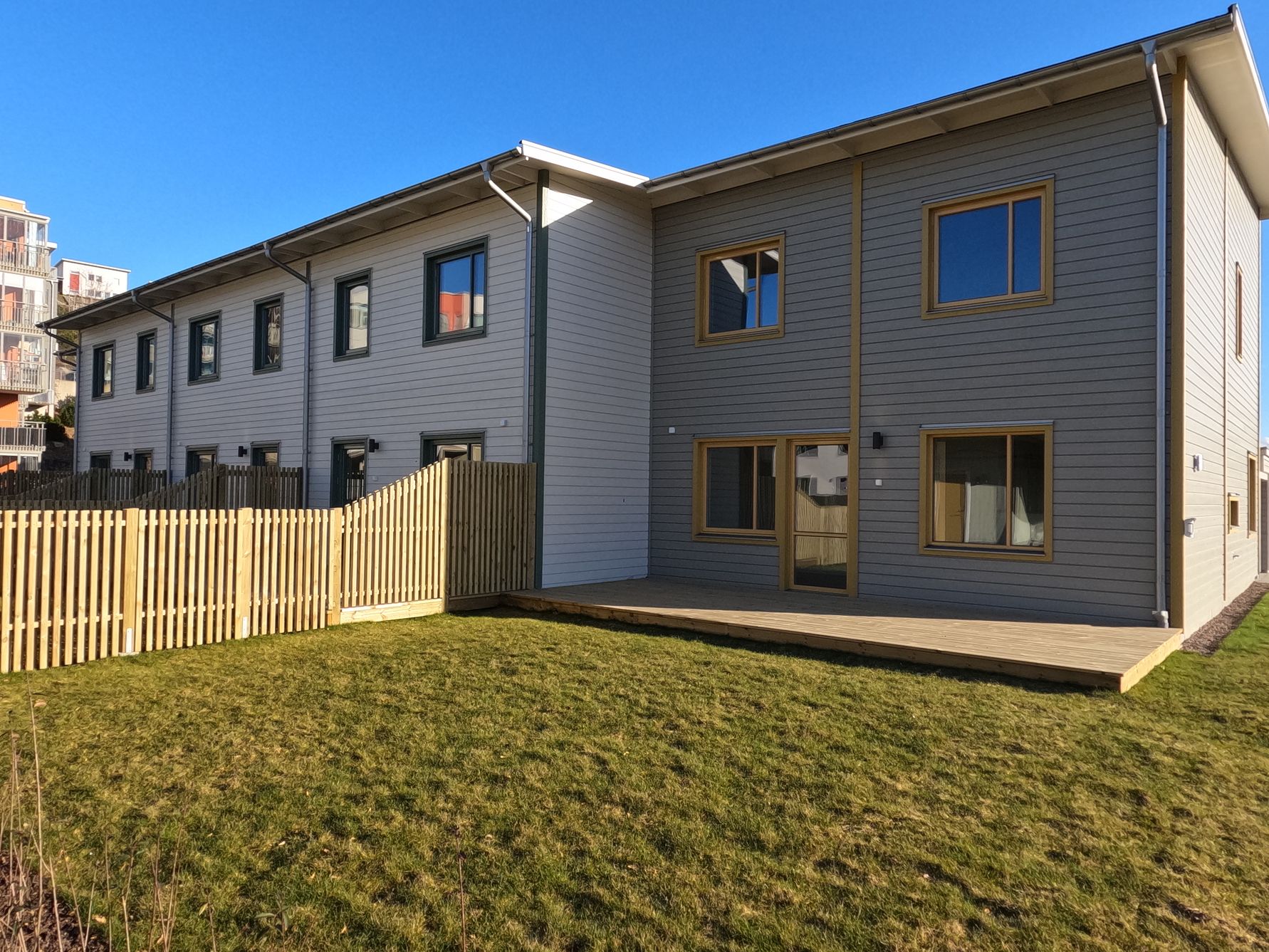 Nybyggda radhus i två våningar med grå fasad, stora fönster och en gräsmatta med trästaket under klarblå himmel