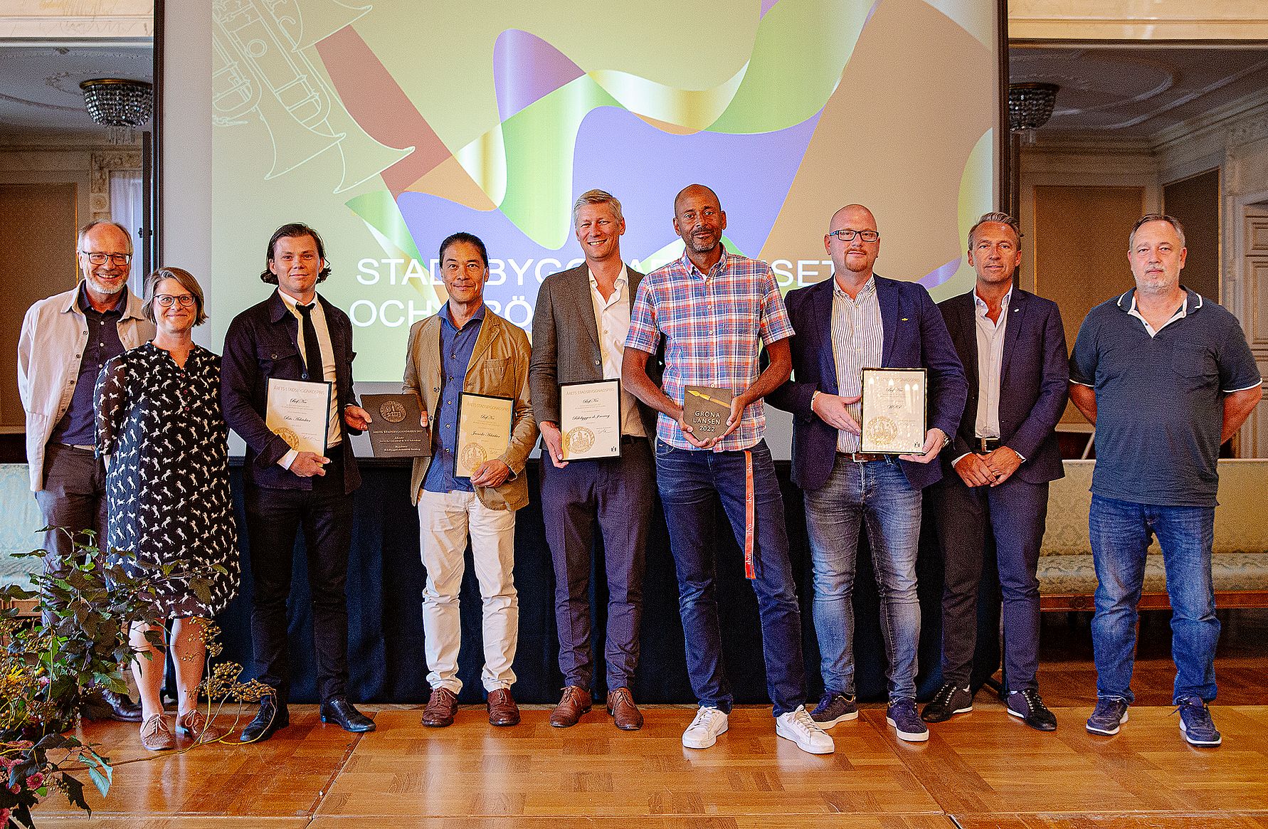 Grupp av åtta personer står på en scen i en sal, flera håller diplom och utmärkelser. Bakgrunden visar en presentationsskärm med färggrafik