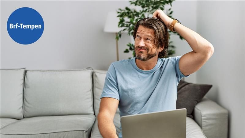 Man i ljusblå t-shirt sitter på en soffa och håller sig på huvudet, med en bärbar dator i knät och en grön växt samt en vit vägglampa i bakgrunden