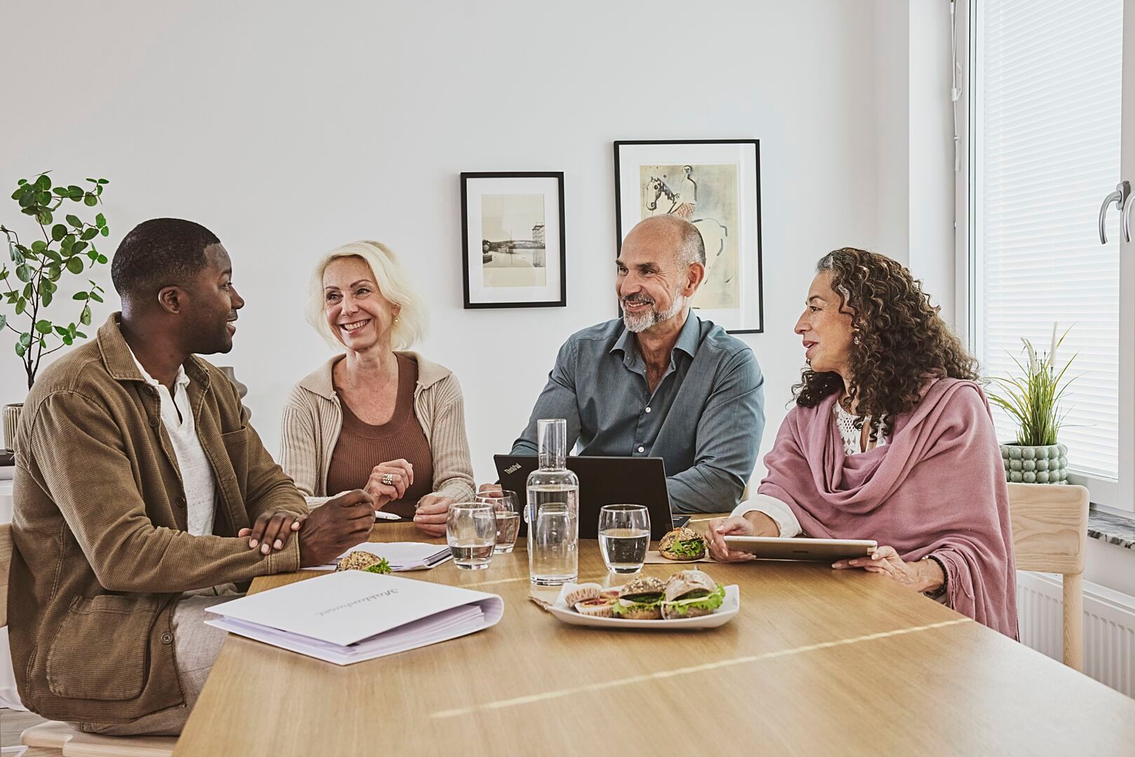 Fyra personer sitter vid ett bord i ett modernt konferensrum med dokument, surfplattor och mat framför sig. Bordet är dekorerat med vattenkaraffer och växtdetaljer
