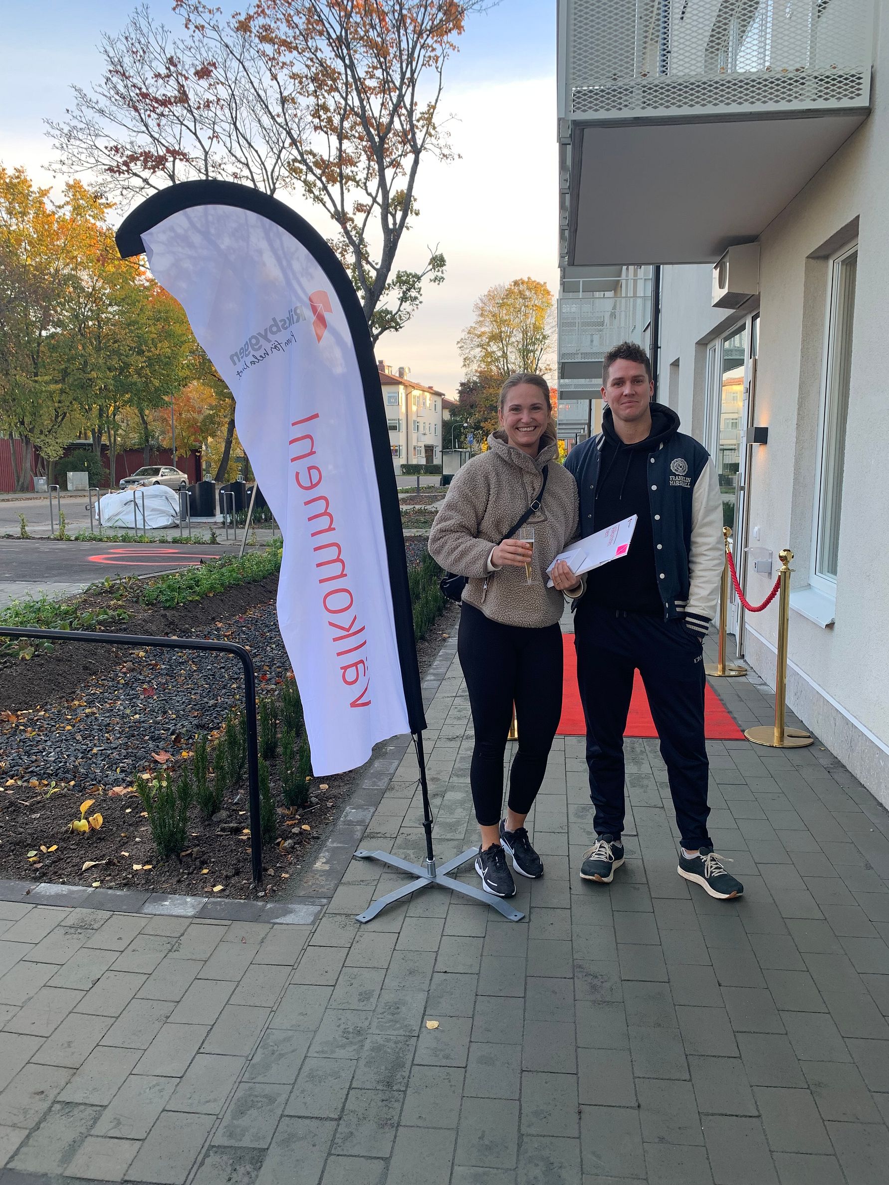 Två personer står på en trottoar bredvid en flagga med texten "Mäklarringen". En röd matta leder in i en byggnad i bakgrunden, med höstlöv på gatan och träd i omgivningen