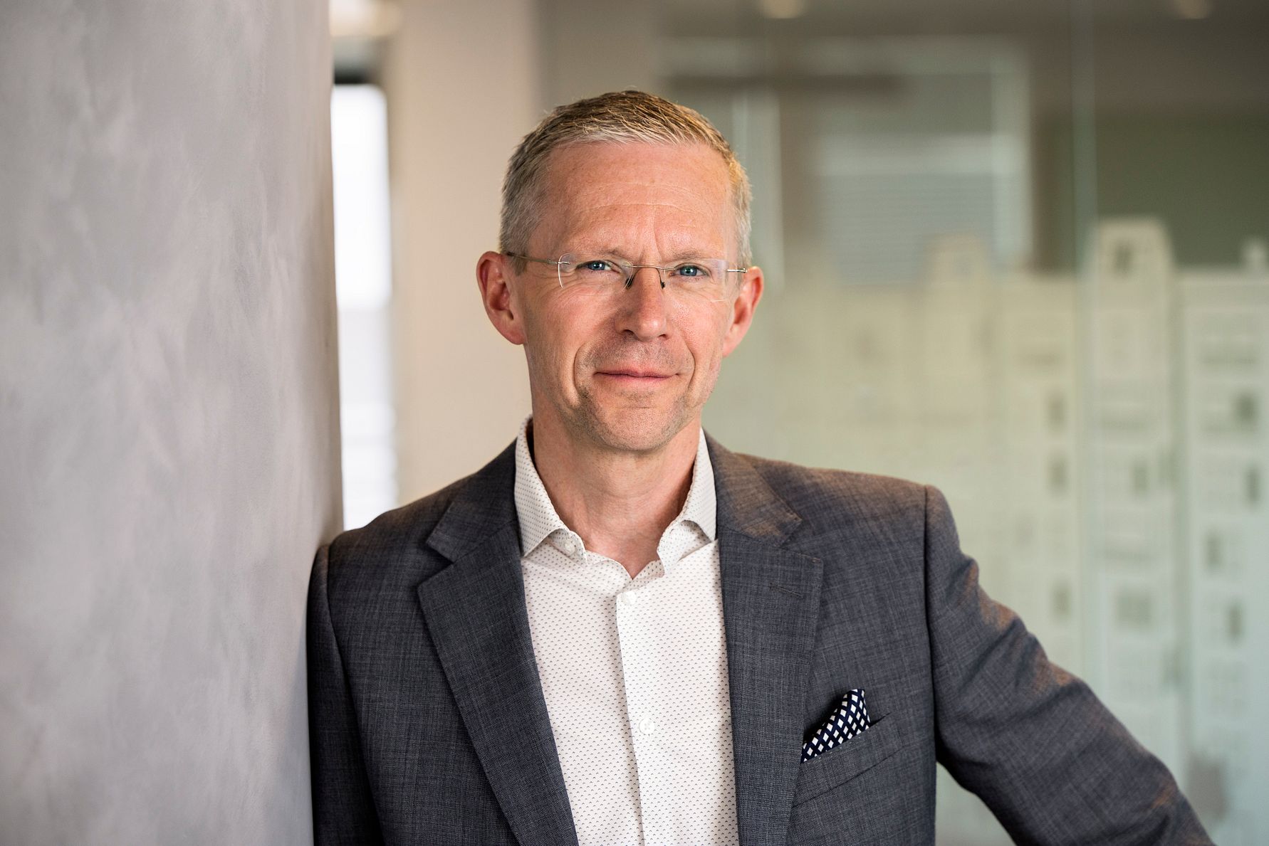 Man iklädd grå kostym och vit skjorta med en prickig näsduk i bröstfickan, stående vid en vägg i en kontorsmiljö