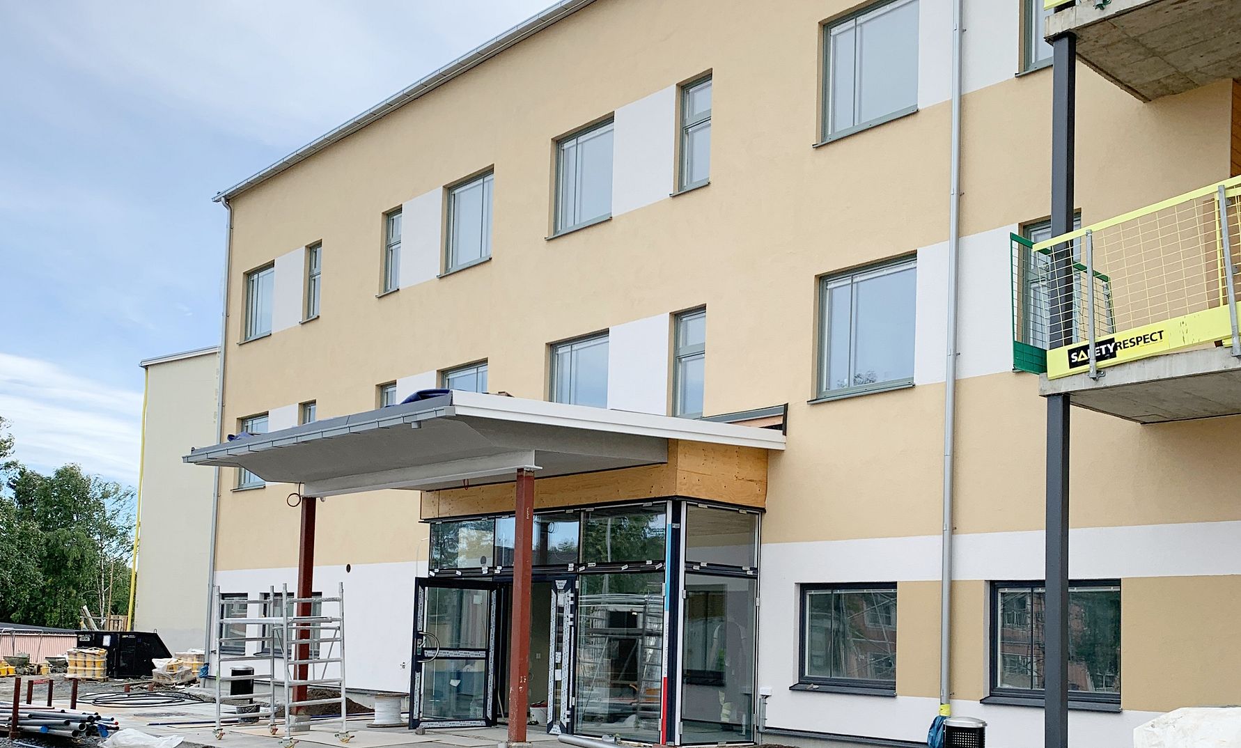 Byggnad med beige fasad och flera fönster, entré med glasdörrar och en överhängande takstruktur