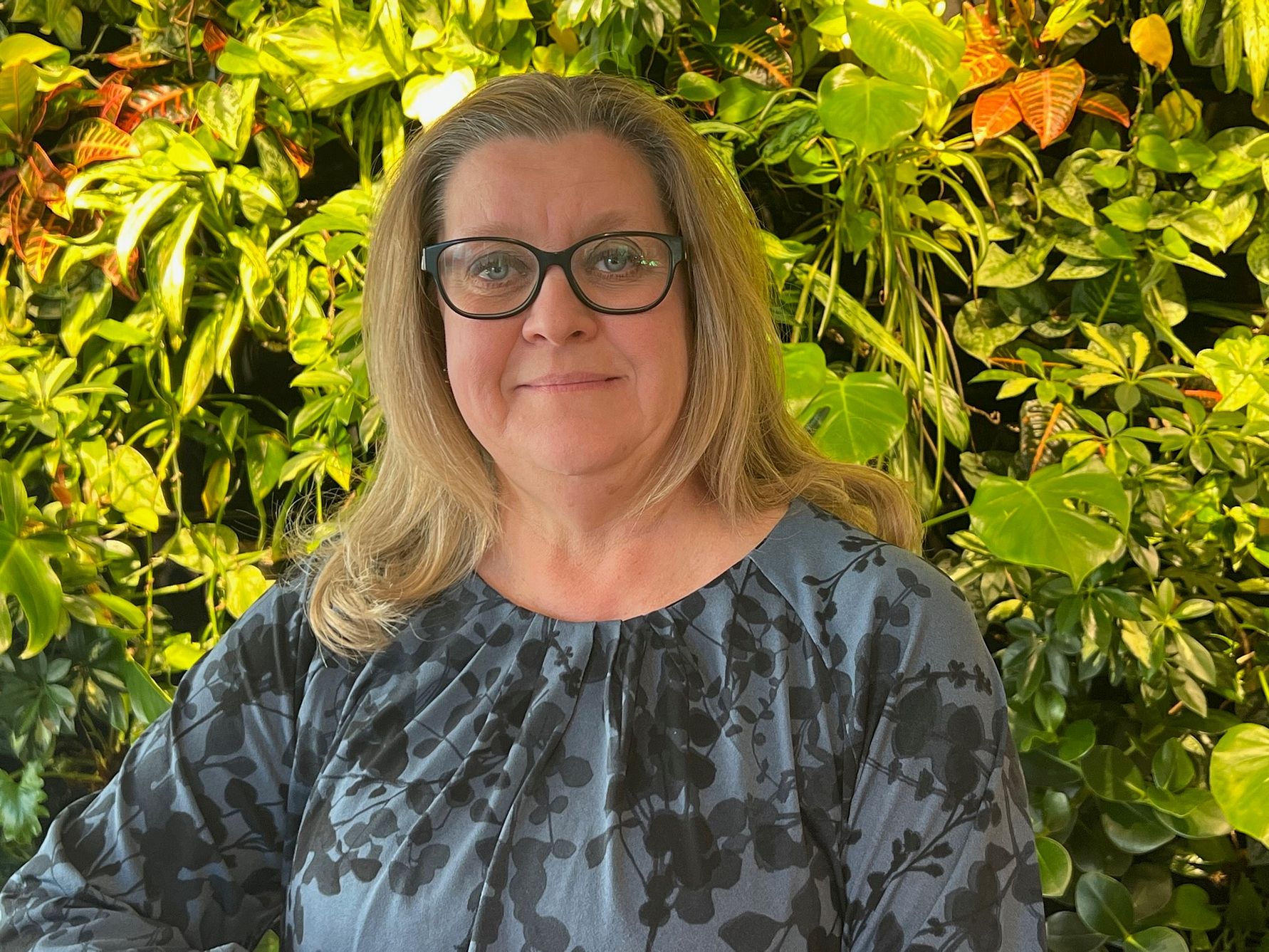 Kvinna i blå blus med blommönster framför en bakgrund av gröna växter