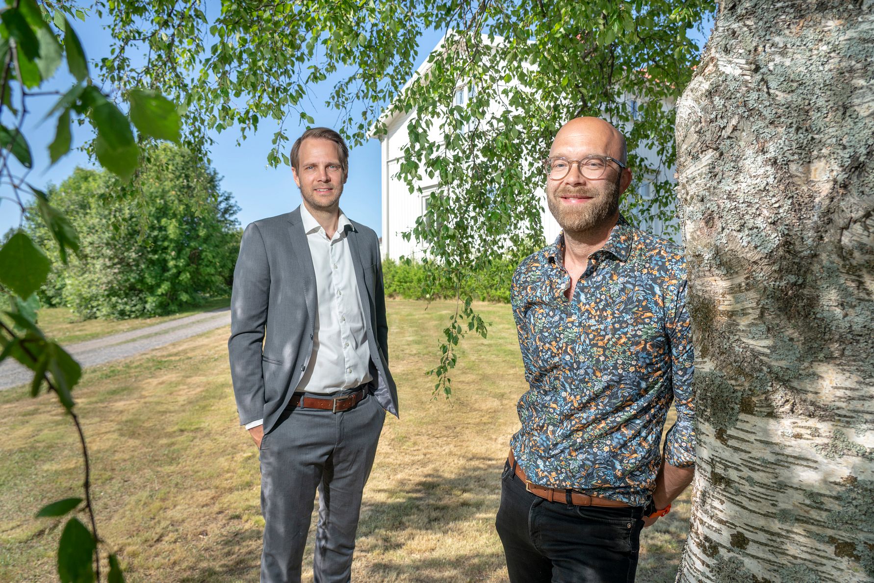Två personer står utomhus på en gräsmatta nära ett träd, med en vit byggnad och träd i bakgrunden under en klarblå himmel
