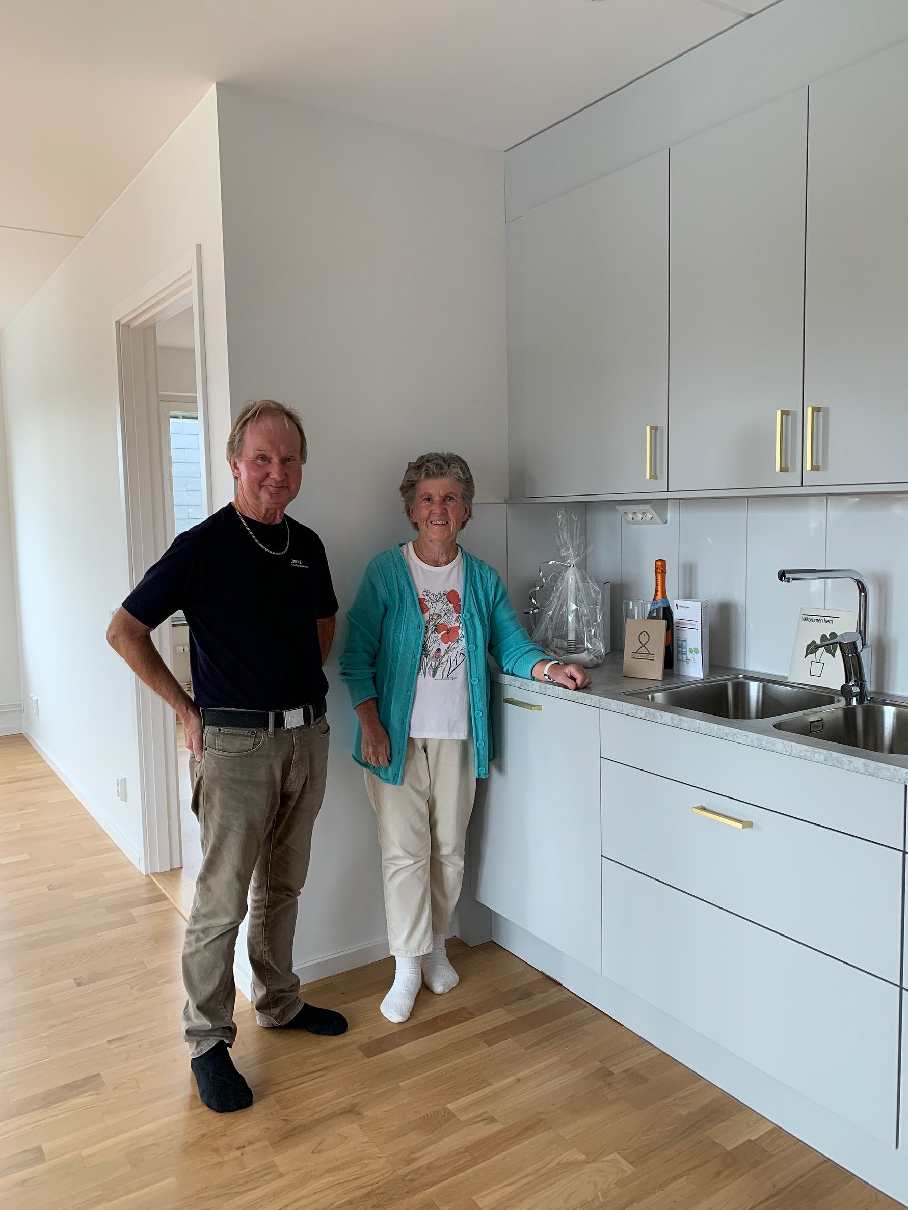 Två personer står i ett modernt kök med ljusa skåpluckor, diskbänk i rostfritt stål och trägolv. En flaska och en presentpåse står på köksbänken