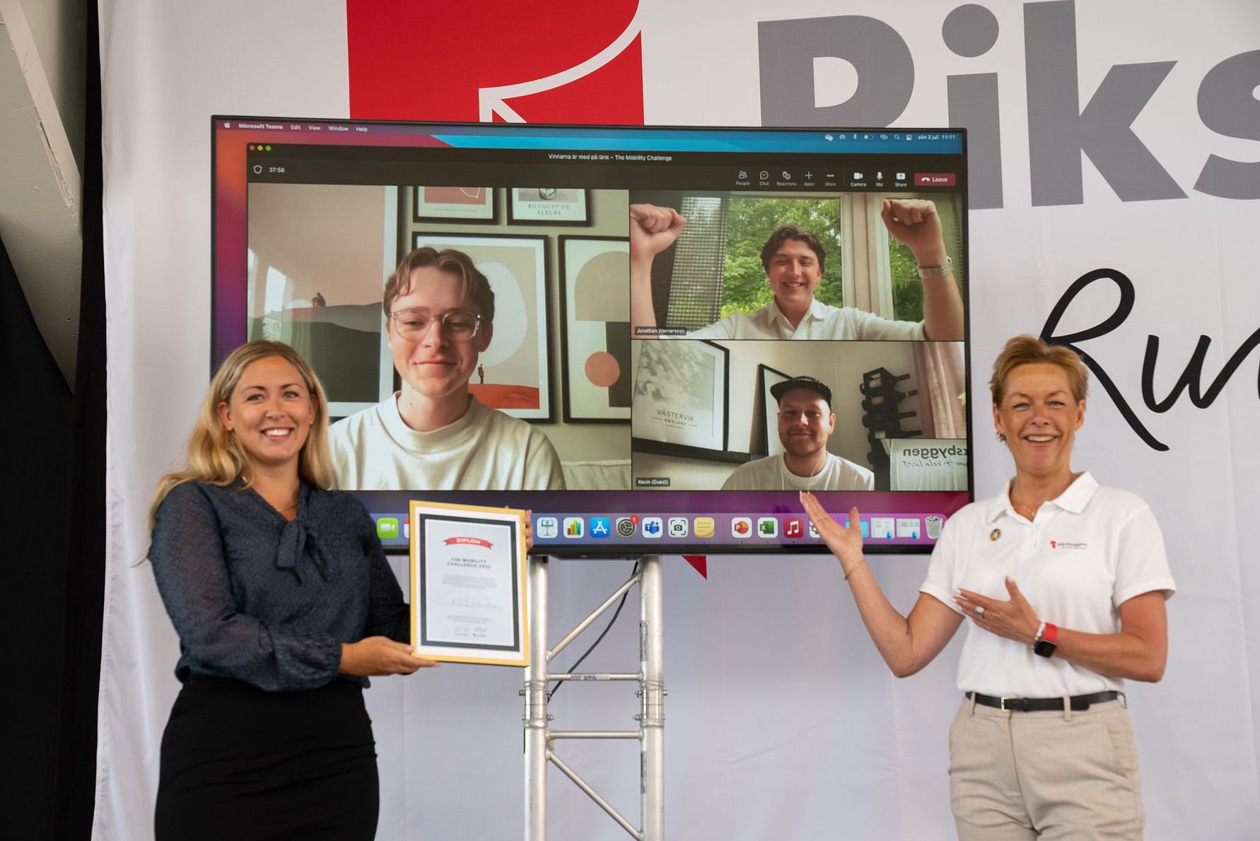 Två personer står framför en stor skärm som visar ett videomöte med fyra deltagare. En av personerna håller ett diplom och den andra pekar mot diplomet, medan en logotyp och text syns i bakgrunden