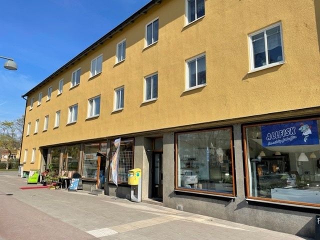 Gul byggnad med butikslokaler i markplan, inklusive en butik med skylt "Allfisk" och ett postombud med gula brevlådor. Klarblå himmel i bakgrunden
