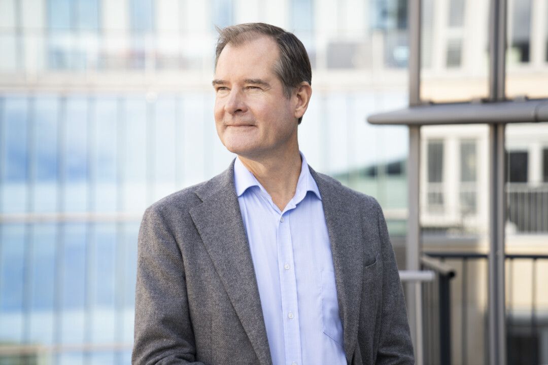 Man i grå kavaj och ljusblå skjorta står framför en modern byggnad med glasfasad