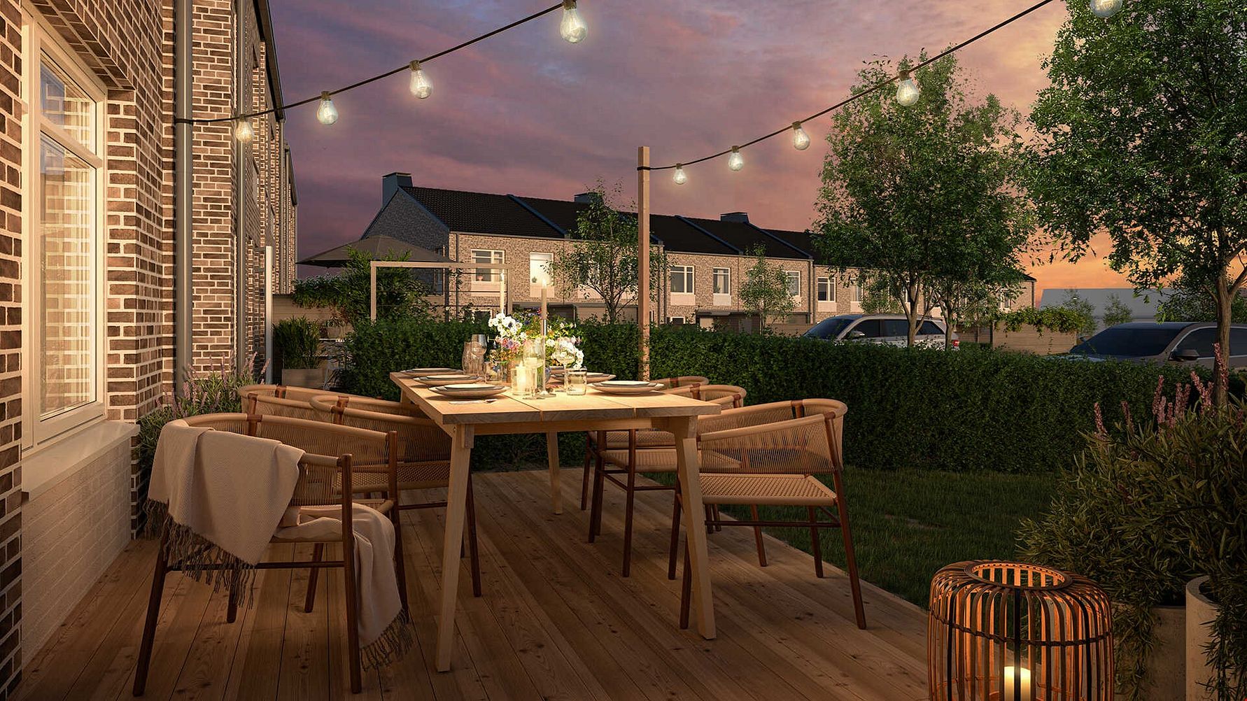 Uteplats med träbord och stolar, dekorerad med ljusslingor och en lykta, omgivet av grönska och bostadshus i bakgrunden under en rosa solnedgång