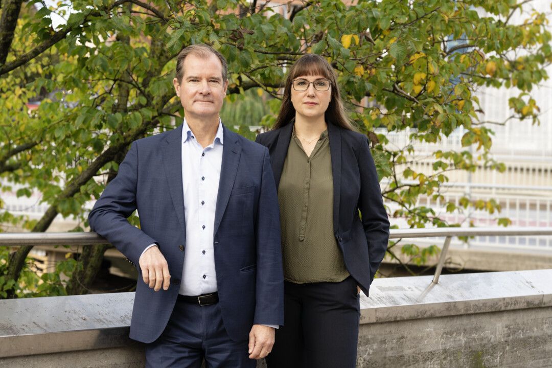 Två personer i formella kläder står utomhus framför ett träd med gröna och gula blad, nära ett räcke