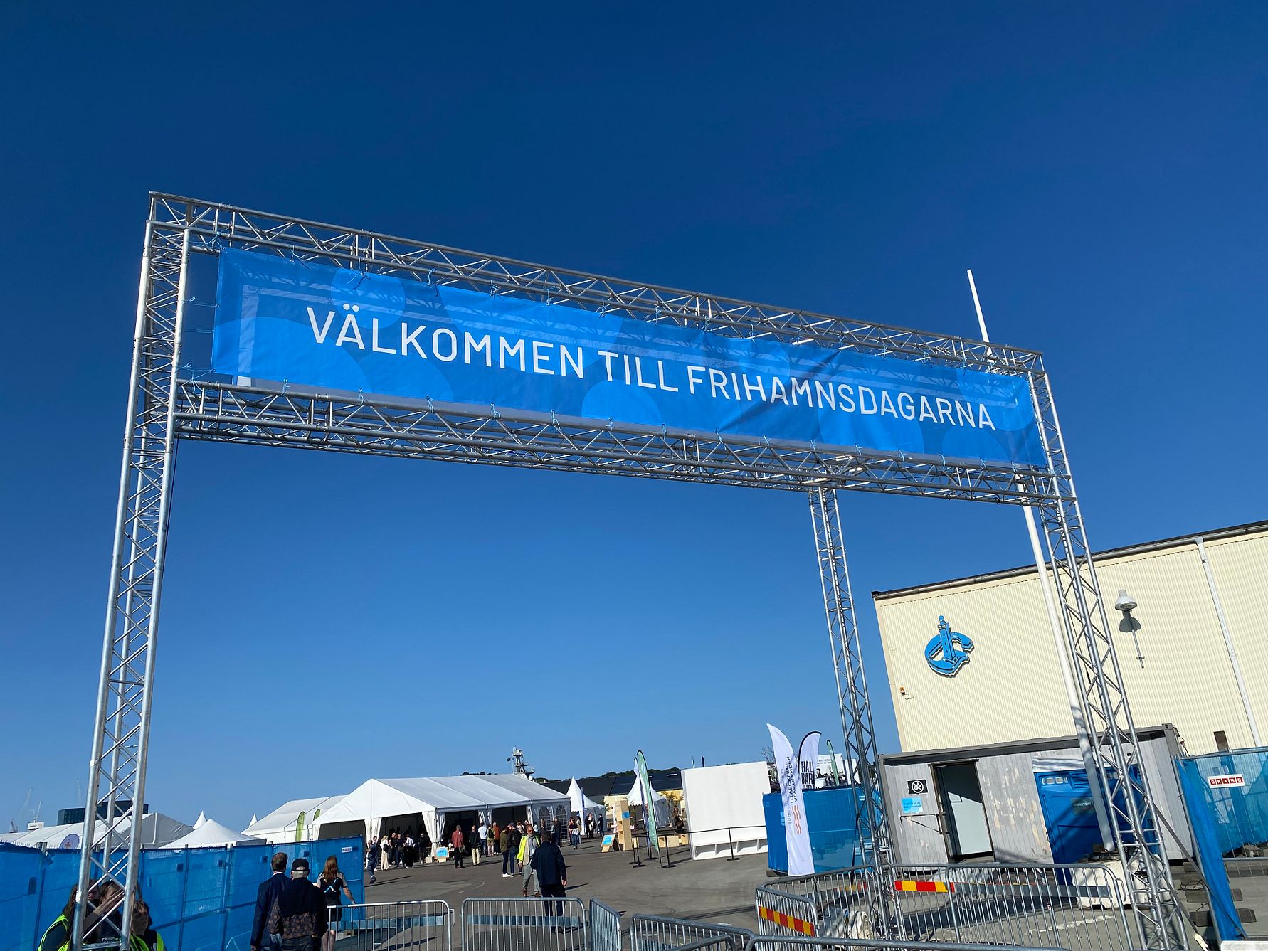 Blått skylt på en metallställning med texten "Välkommen till Frihamnsdagarna" mot en klarblå himmel, omgiven av tält och byggnader