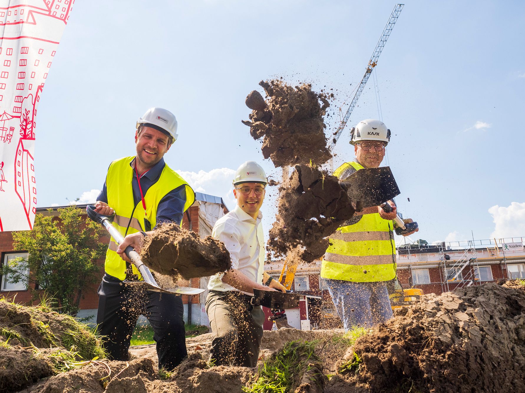 Byggarbetare med hjälmar och varselkläder som utför grävarbete på en byggarbetsplats, med en kran och byggnader i bakgrunden