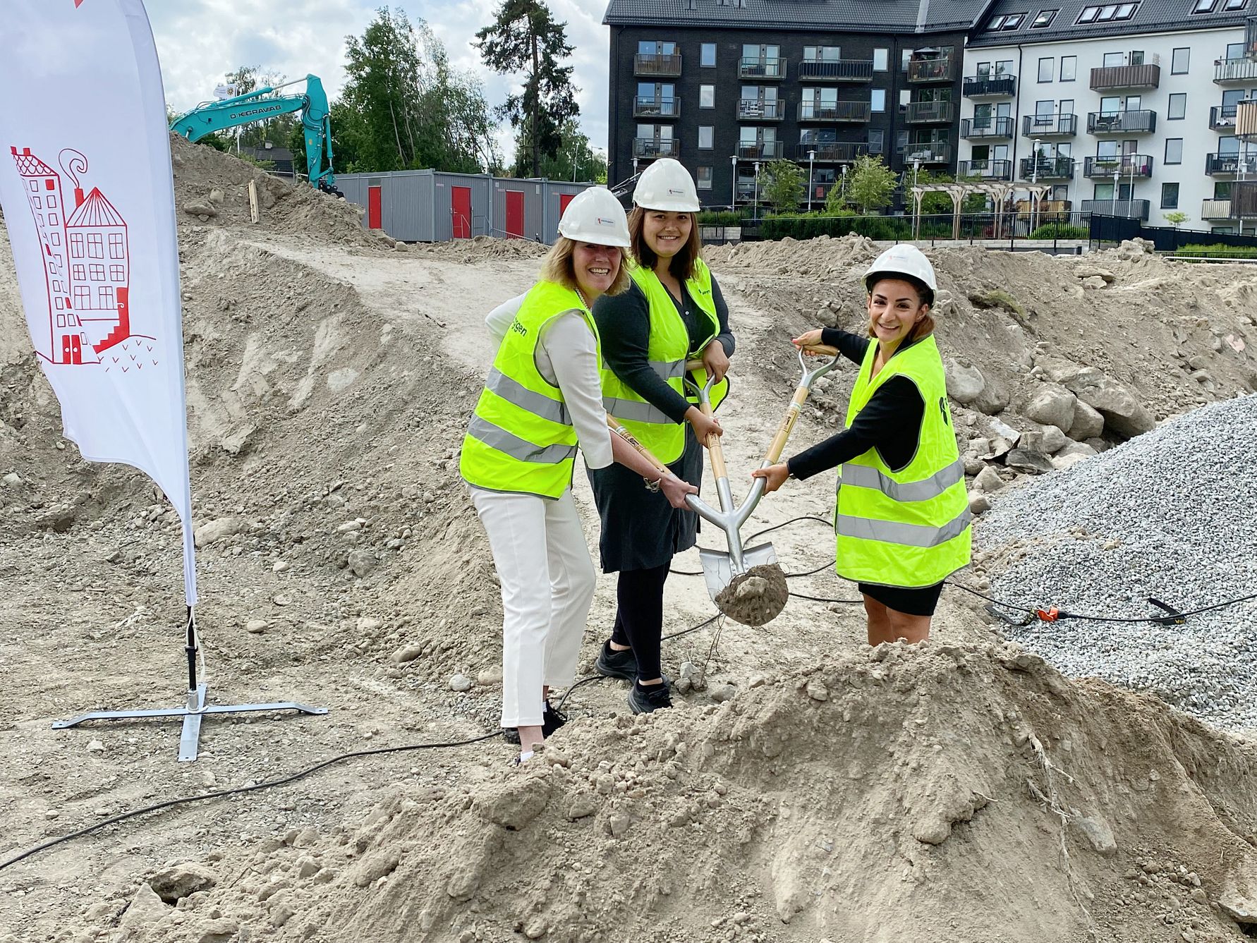 Fyra personer i reflexvästar och hjälmar deltar i en ceremonisk spadtagning på en byggarbetsplats, med högar av jord och en byggnad i bakgrunden