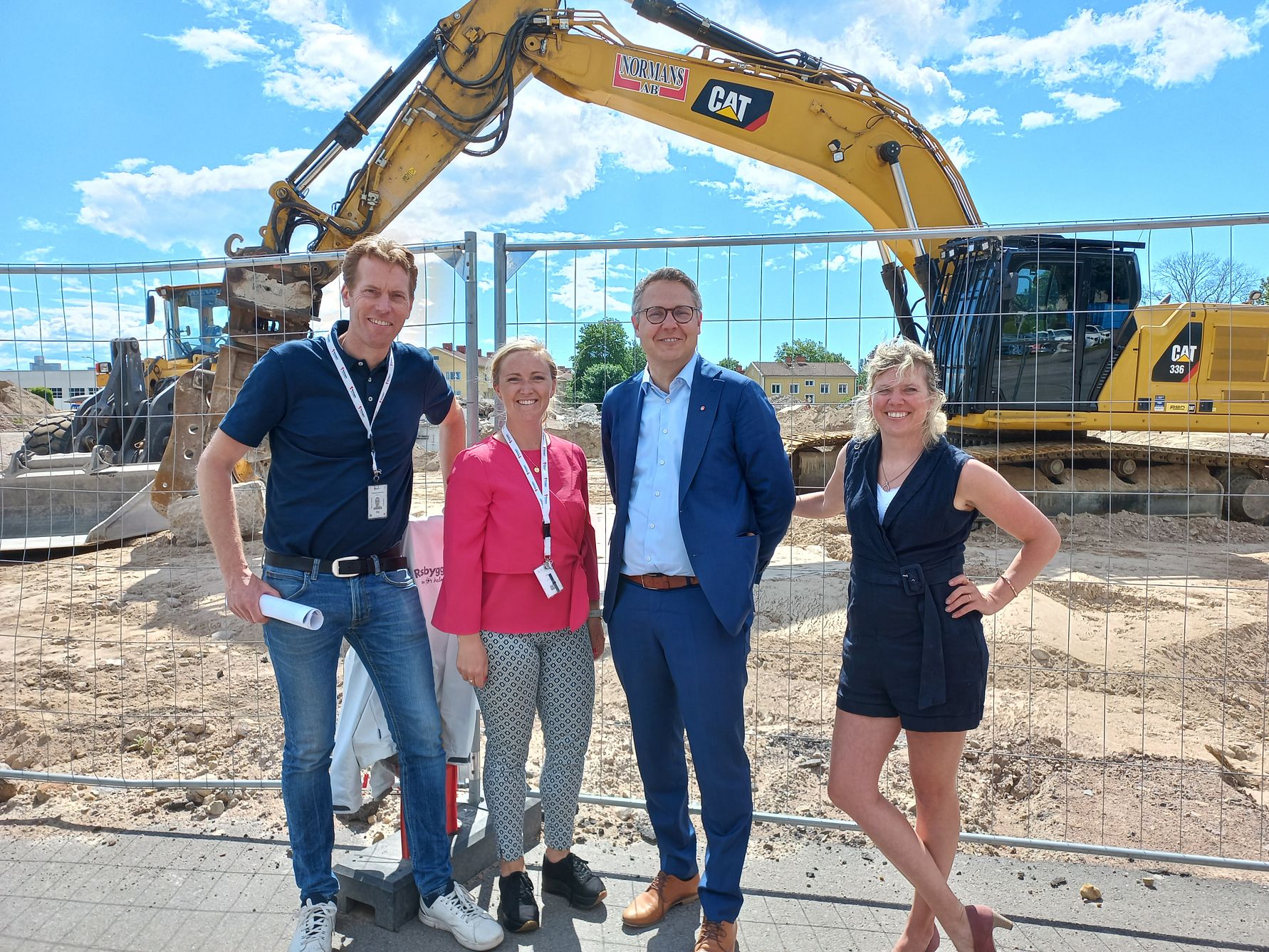 Fyra personer står framför ett grävmaskinområde, med ett stängsel och en Caterpillar-grävmaskin i bakgrunden, under en klarblå himmel