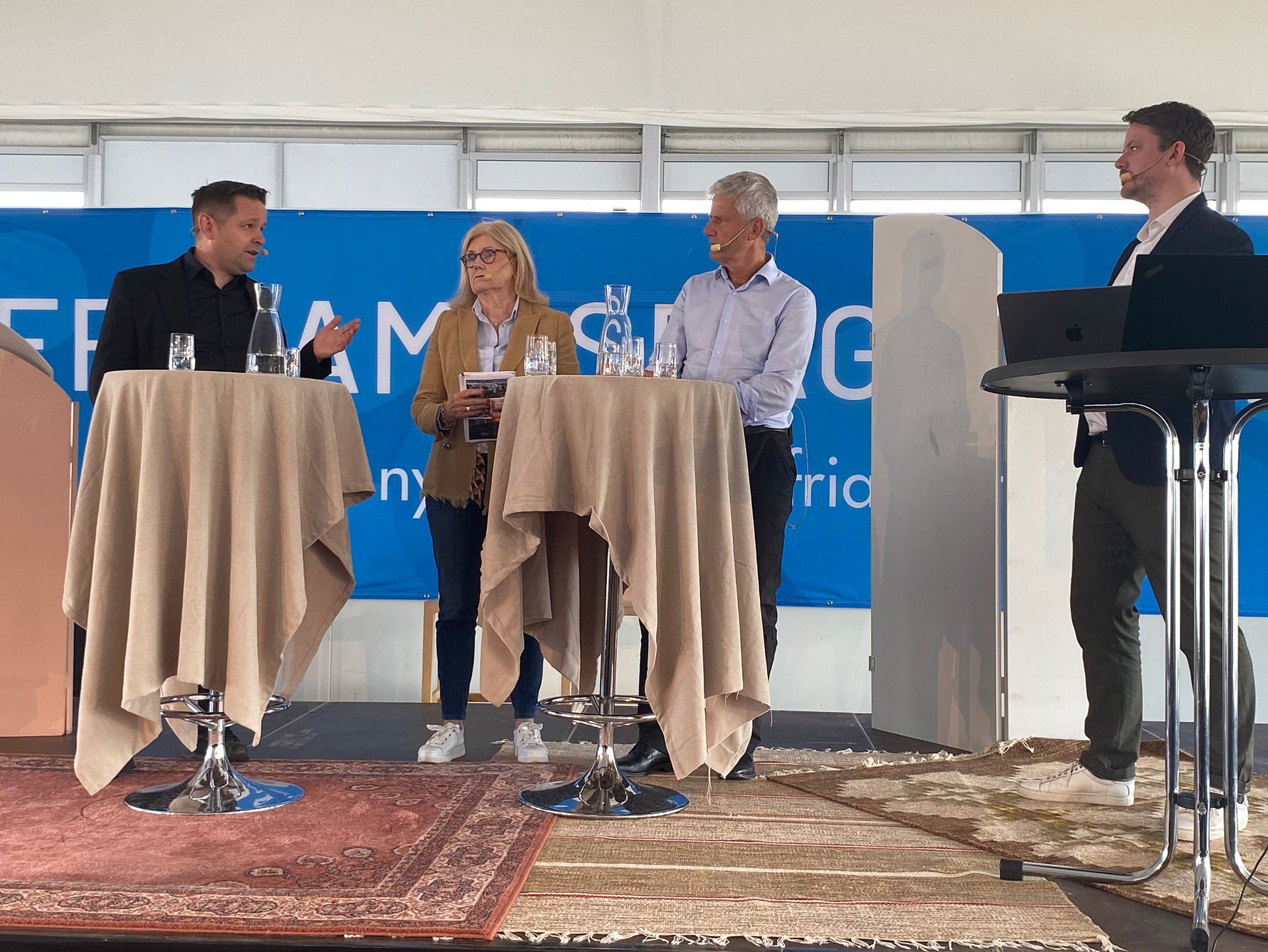 Fyra personer står på en scen framför en blå bakgrund, med två höga bord täckta med beige dukar och dryckesglas