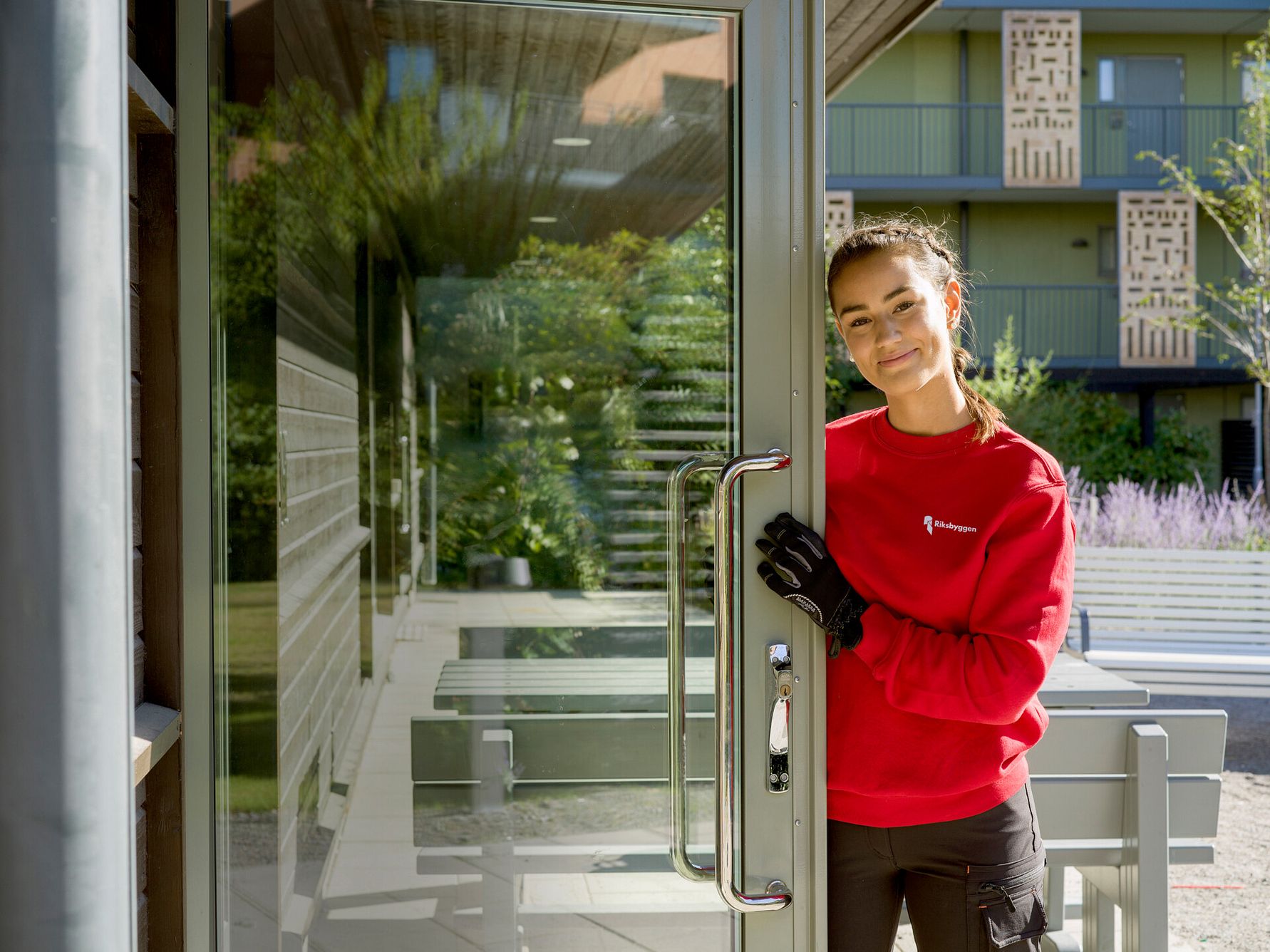 En person i röd tröja och svarta handskar öppnar en glasdörr, med moderna byggnader och gröna växter i bakgrunden