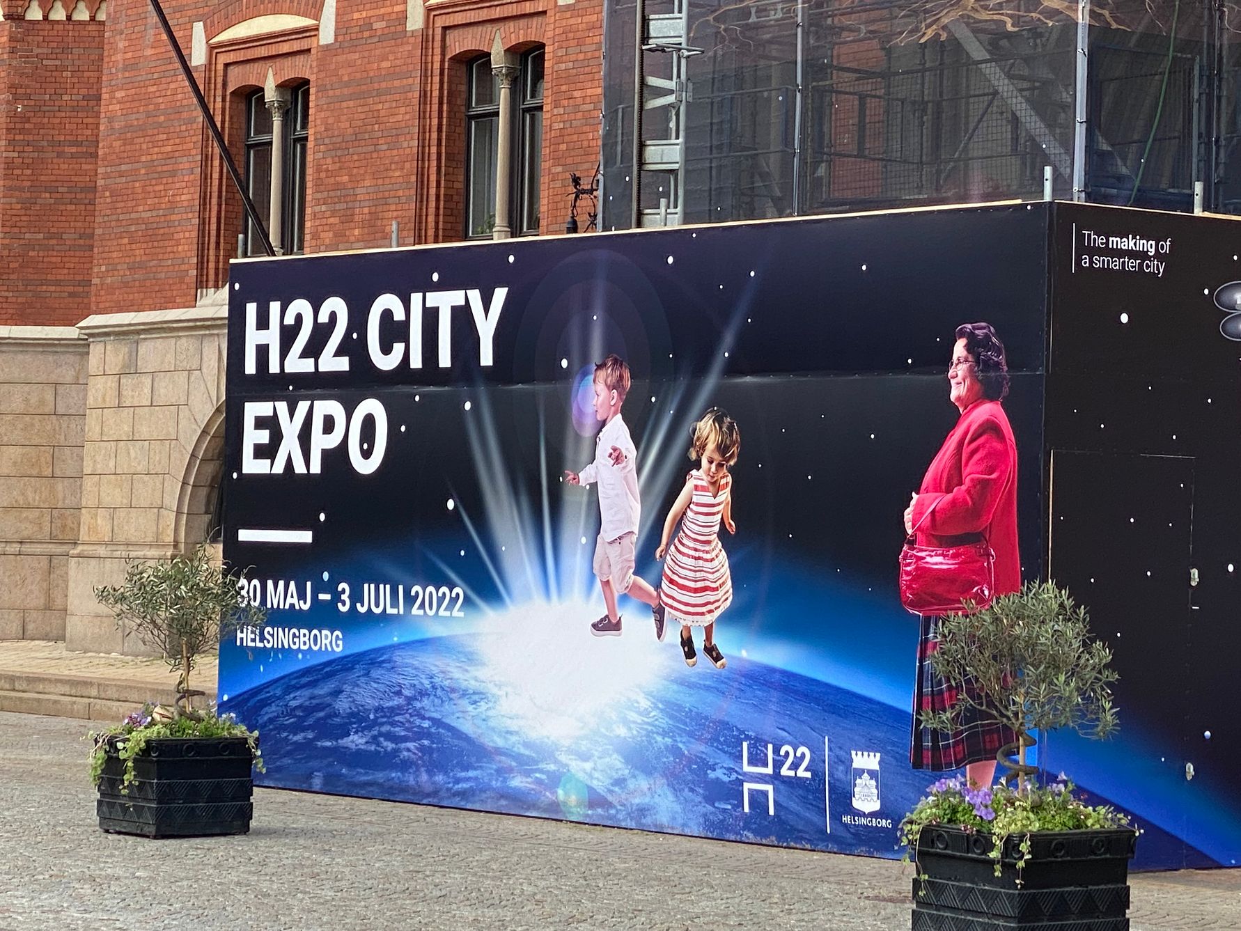 Reklamskylt för H22 City Expo i Helsingborg, med text och färgglada bilder av två barn som hoppar och en vuxen person på en illustration av jorden, omgiven av byggnader och två planteringar i förgrunden