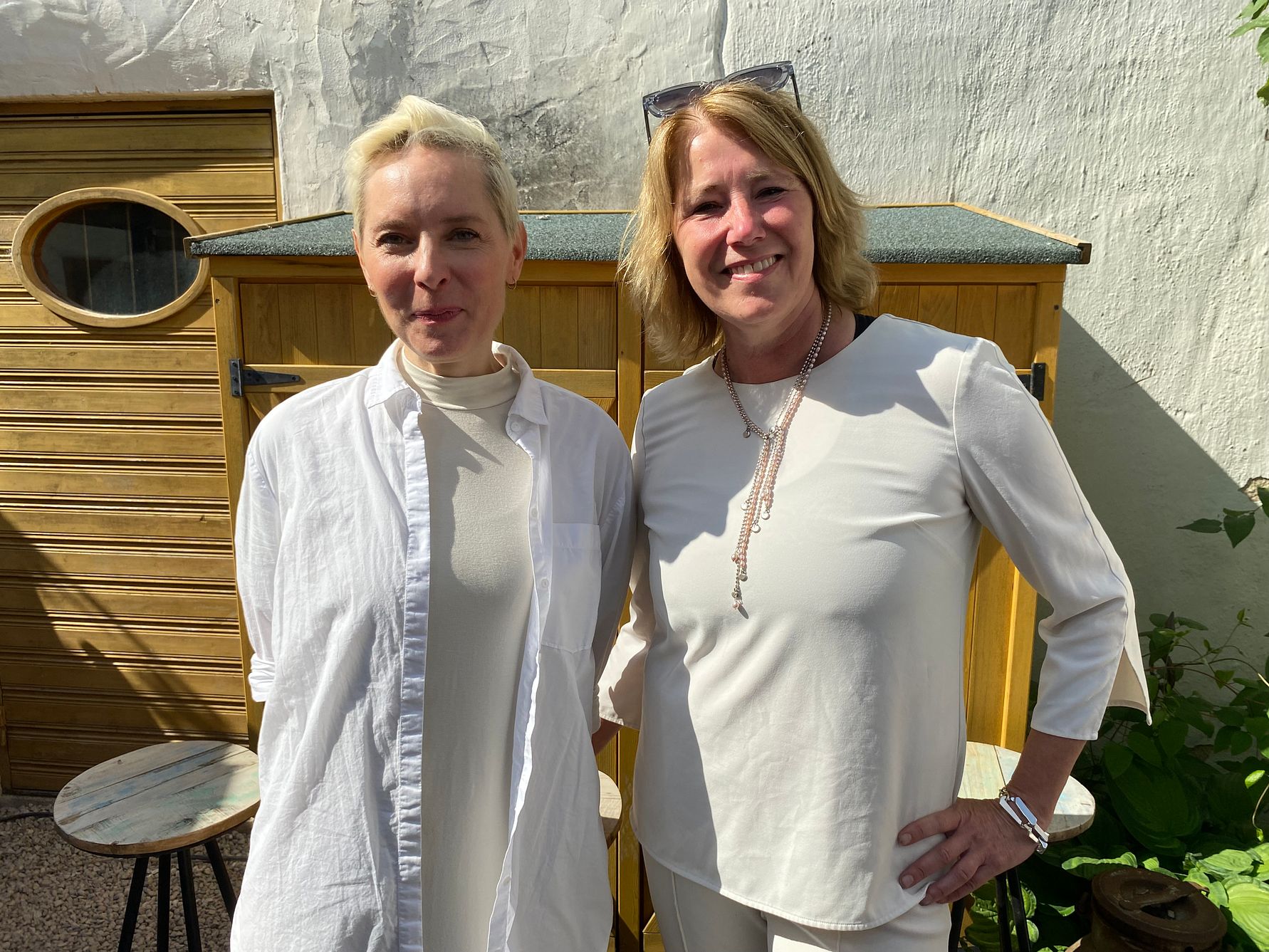 Två personer klädda i ljusa kläder står framför en gul träbyggnad med en rund dörr och växtlighet i bakgrunden