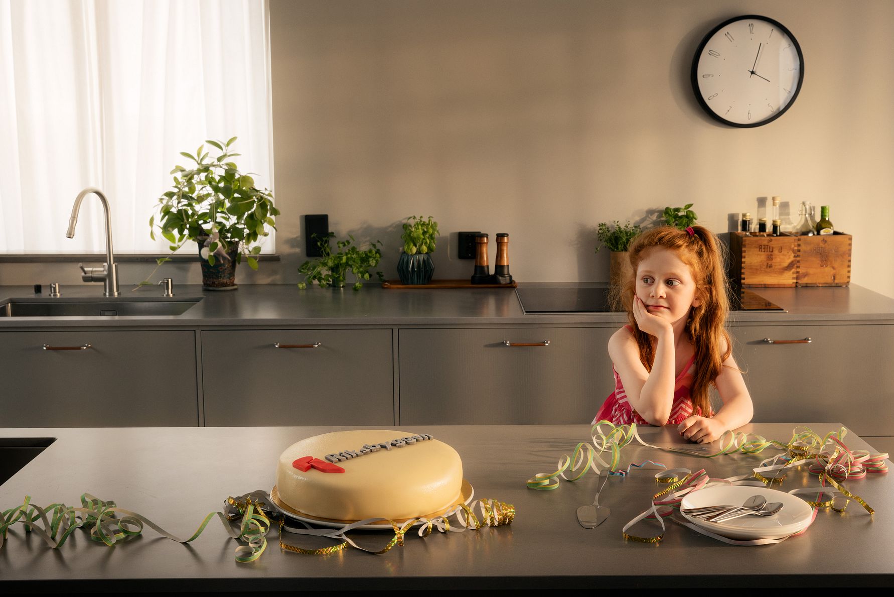 Ett modernt kök med grå arbetsytor, växter i bakgrunden och en vit tårta dekorerad med text och ett rött hjärta på ett bord, omgiven av serpentiner och tallrikar
