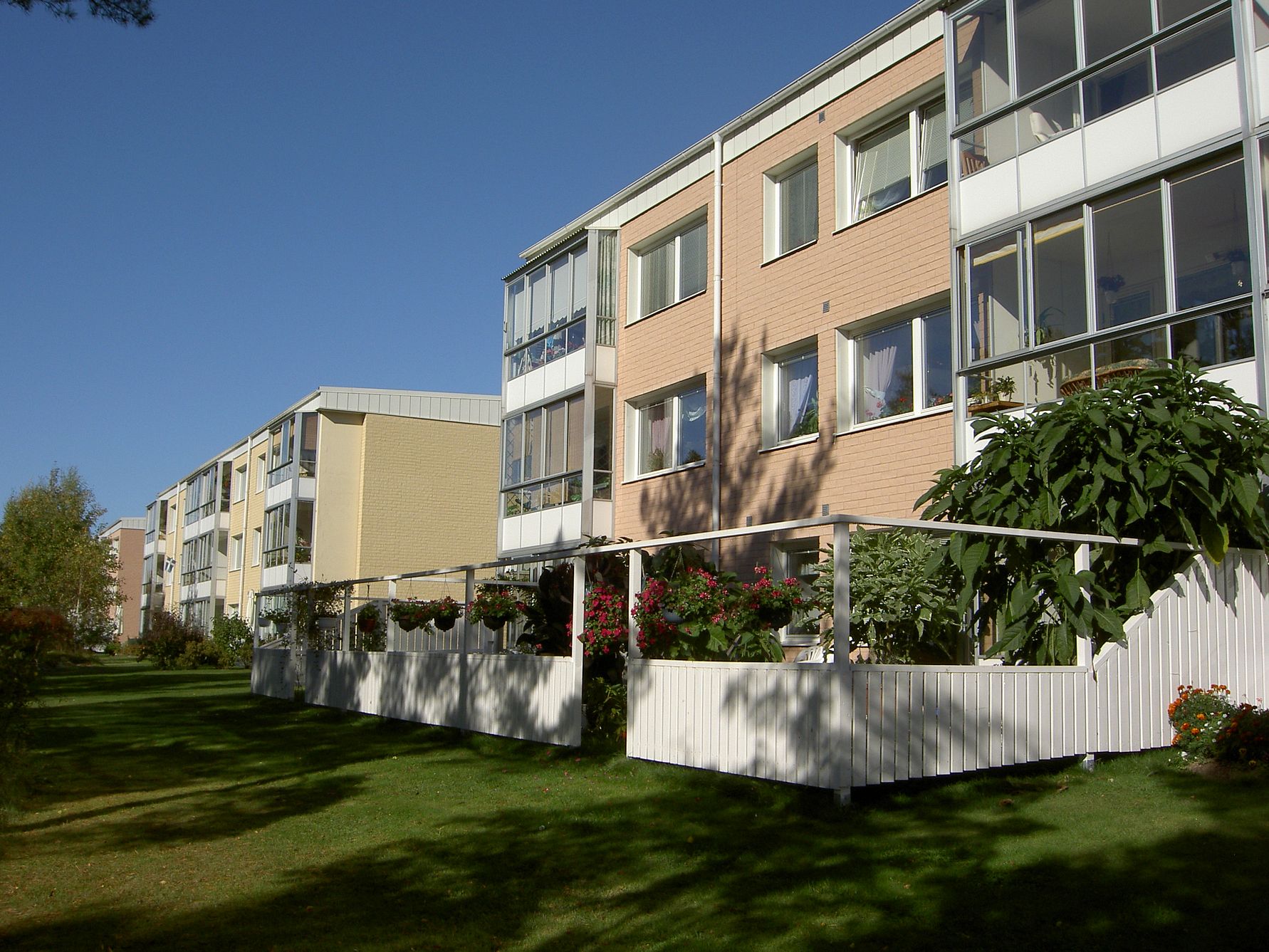 Flerbostadshus med fasader i ljusa färger, balkonger och grönområde med träd och buskar i förgrunden