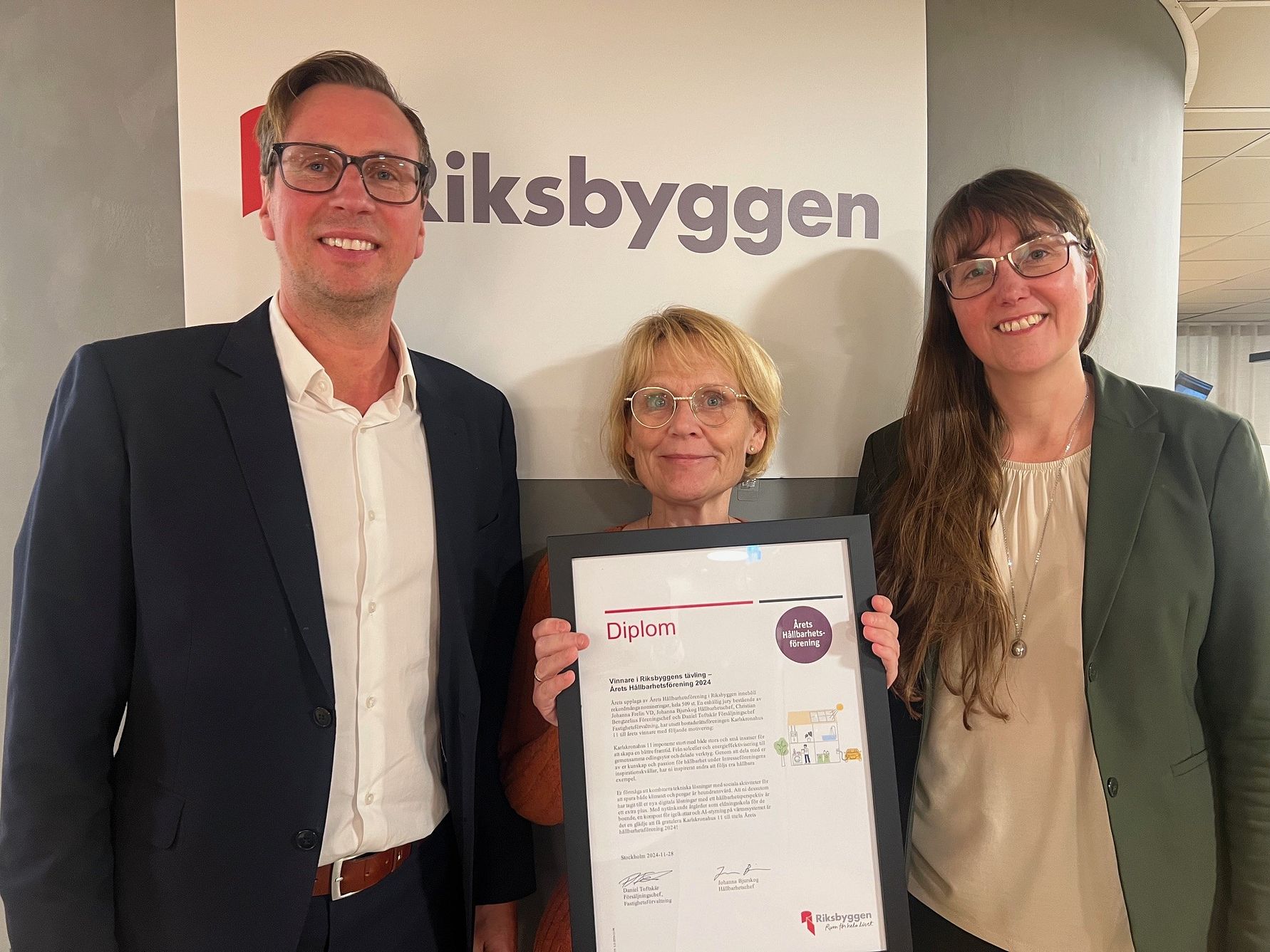 Tre personer står framför en skylt med texten "Riksbyggen" och håller ett diplom