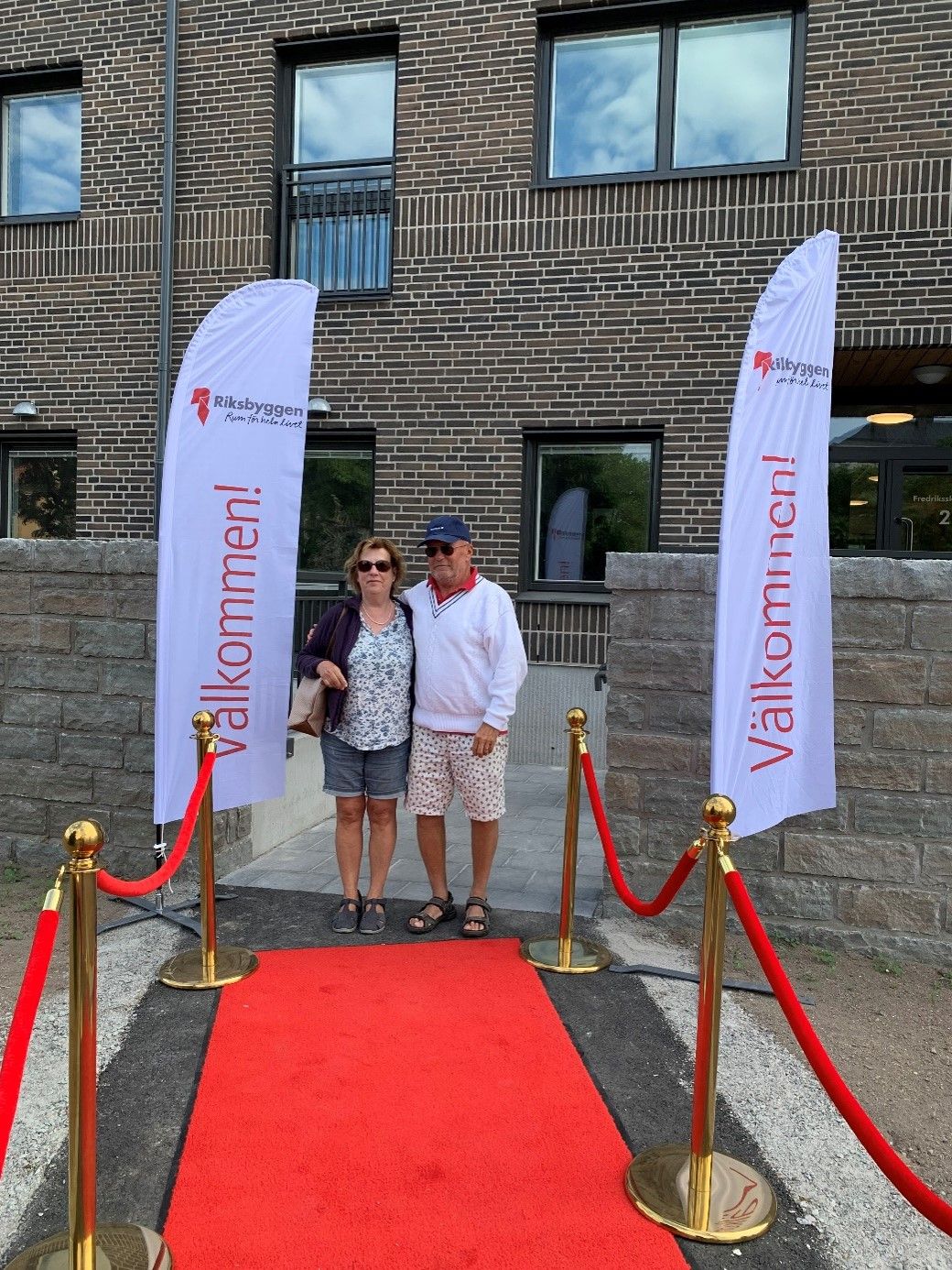 Två personer står på en röd matta omgiven av röda avspärrningsband framför en byggnad med tegelvägg. Två vita flaggor med texten "Välkommen!" är placerade på vardera sidan av mattan
