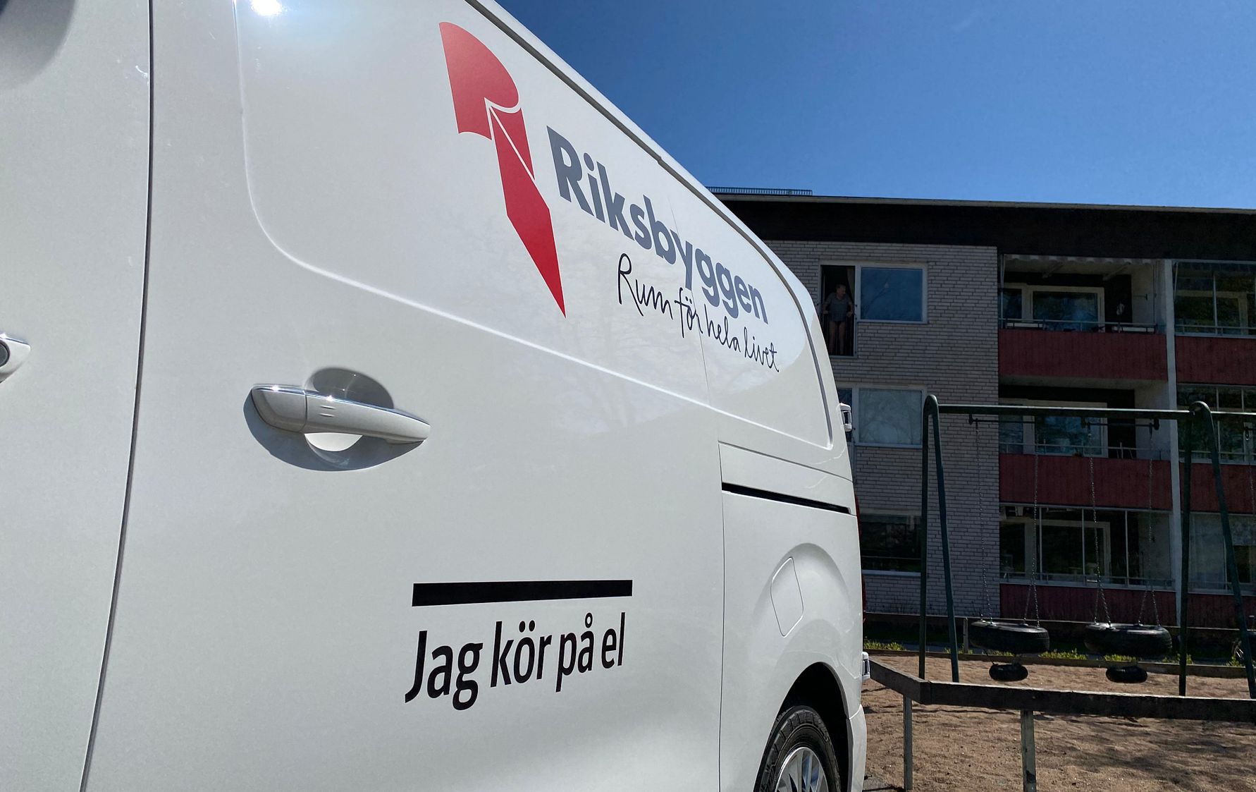 Vit skåpbil med Riksbyggen-logotyp och slogan "Jag kör på el" parkerad framför en byggnad under en klarblå himmel