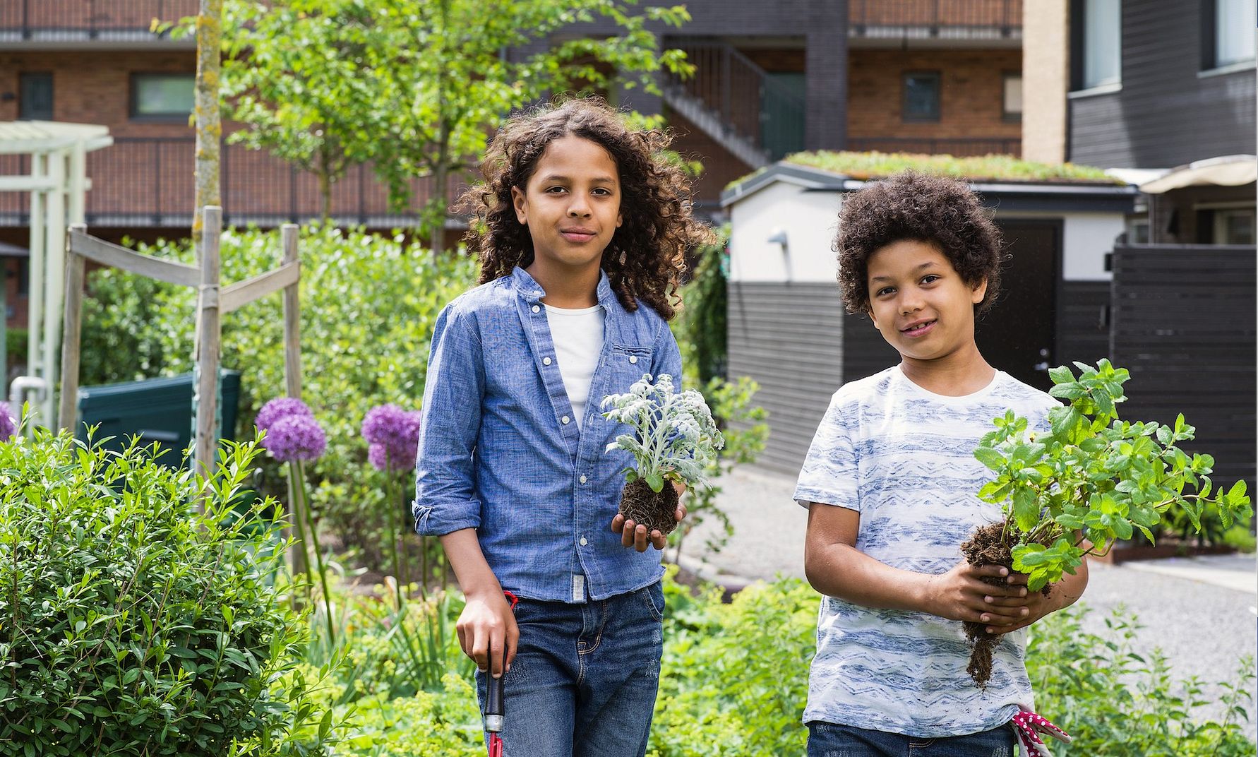 Två personer står i en grönskande trädgård, och håller i varsitt växt med jordiga rötter. Bakgrunden visar ett bostadsområde med byggnader och växande växter