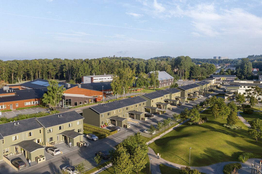 Flygvy över ett bostadsområde med radhus, gröna gräsmattor och omgivande träd, med byggnader och skog i bakgrunden
